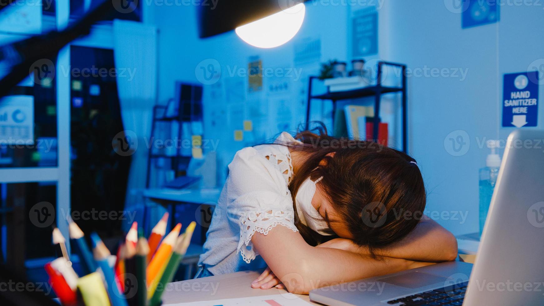 Freelance Asia exhausted lady wear face mask sleeping at new normal home office. Working from house overload at night, Remotely, self isolation, social distance, quarantine for corona virus prevention photo