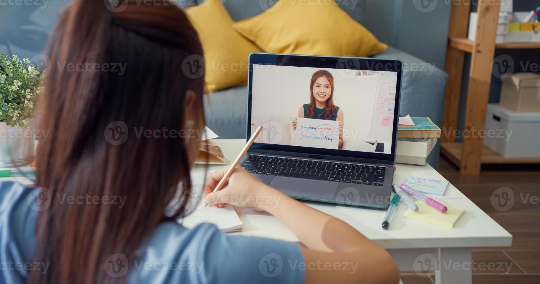 joven asiática con videollamada de computadora portátil de uso casual aprender en línea con el maestro escribir conferencia cuaderno sala de estar en casa. Aislar el concepto de pandemia de coronavirus de educación en línea e-learning. foto