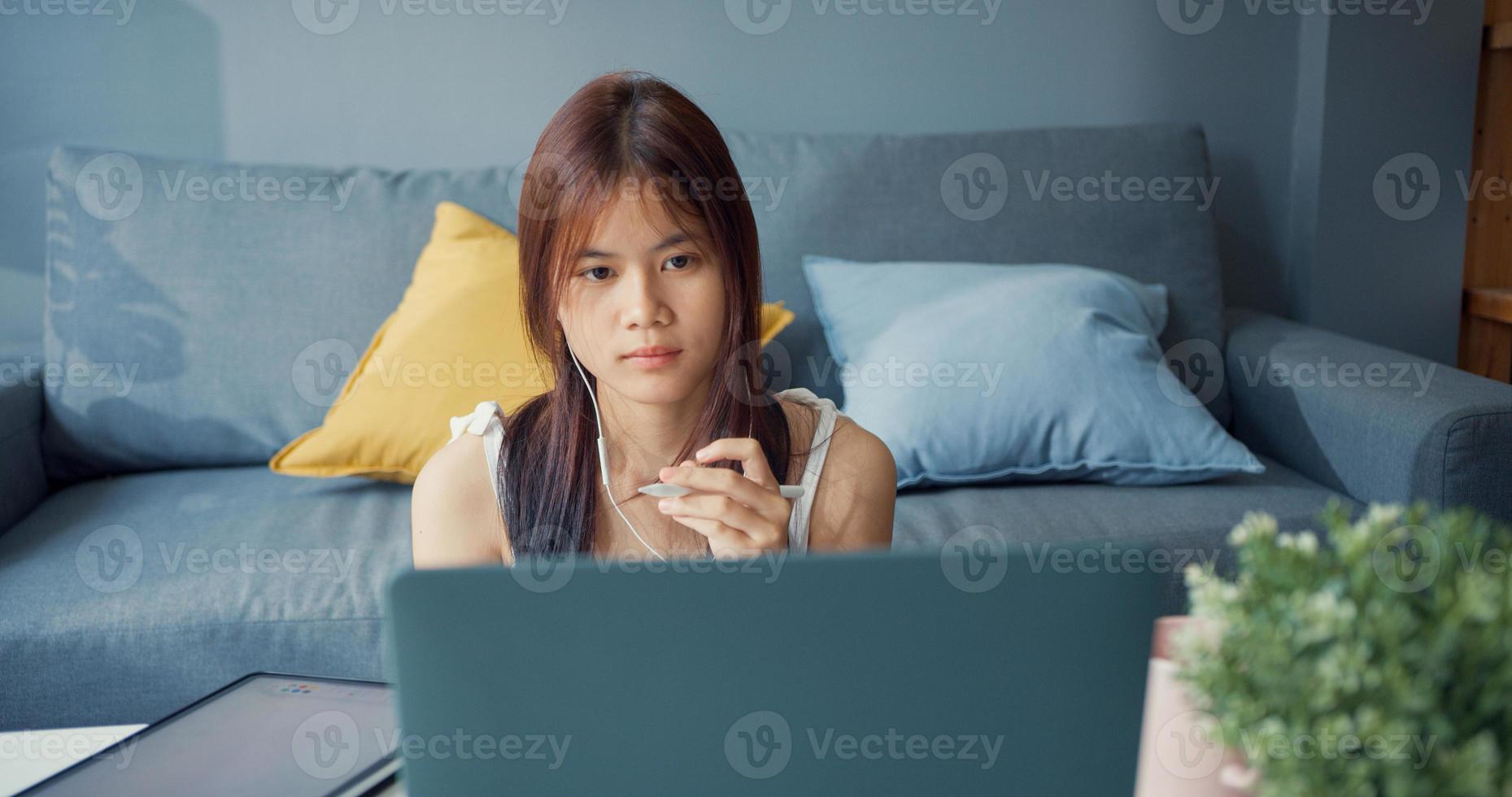 Young Asia girl teenager with casual shirt wear earphone use laptop learn online write lecture in laptop in living room at house. Isolate education online e-learning coronavirus pandemic concept. photo