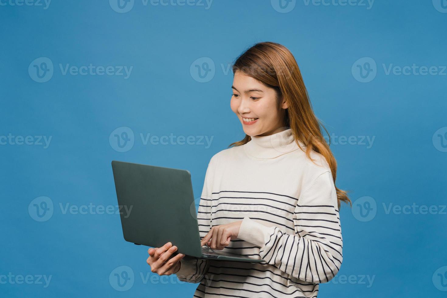 Jovencita asiática usando laptop con expresión positiva, sonríe ampliamente, vestida con ropa casual sintiendo felicidad y parada aislada sobre fondo azul. feliz adorable mujer alegre se regocija con el éxito. foto