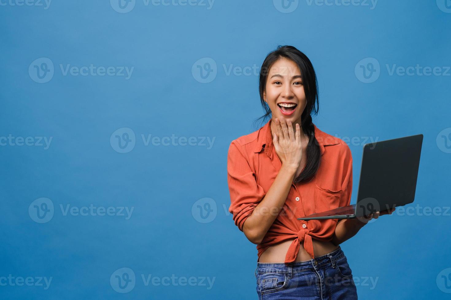 Surprised young Asia lady using laptop with positive expression, smile broadly, dressed in casual clothing and looking at camera on blue background. Happy adorable glad woman rejoices success. photo