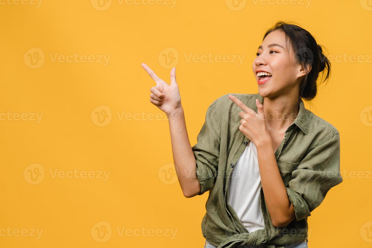 Portrait of young Asian lady smiling with cheerful expression, shows something amazing at blank space in casual clothing and standing isolated over yellow background. Facial expression concept. photo