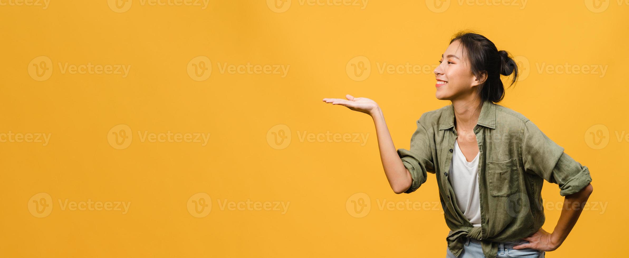 Portrait of young Asian lady smiling with cheerful expression, shows something amazing at blank space in casual clothing and standing isolated over yellow background. Panoramic banner with copy space. photo