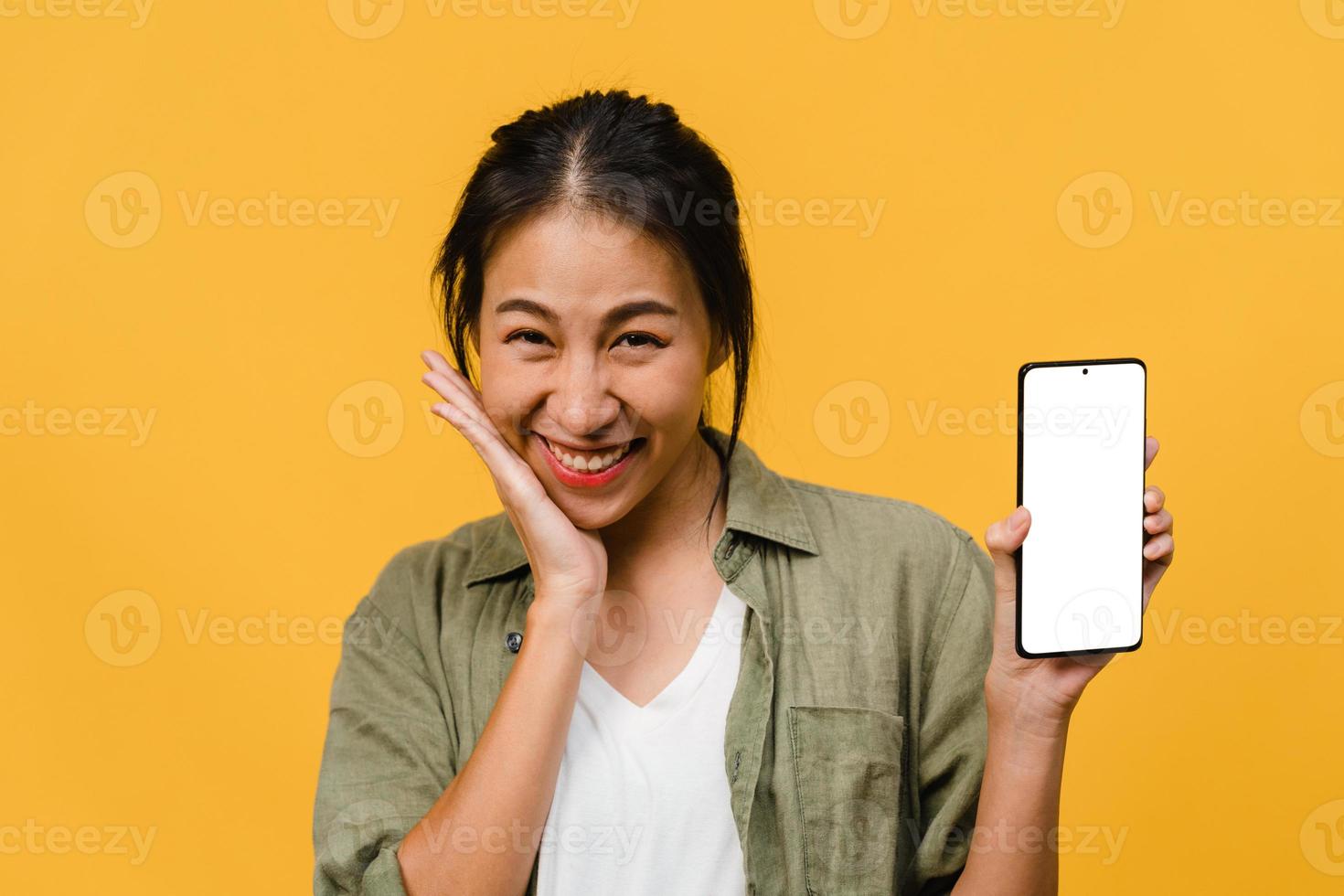 Young Asia lady show empty smartphone screen with positive expression, smiles broadly, dressed in casual clothing feeling happiness on yellow background. Mobile phone with white screen in female hand. photo