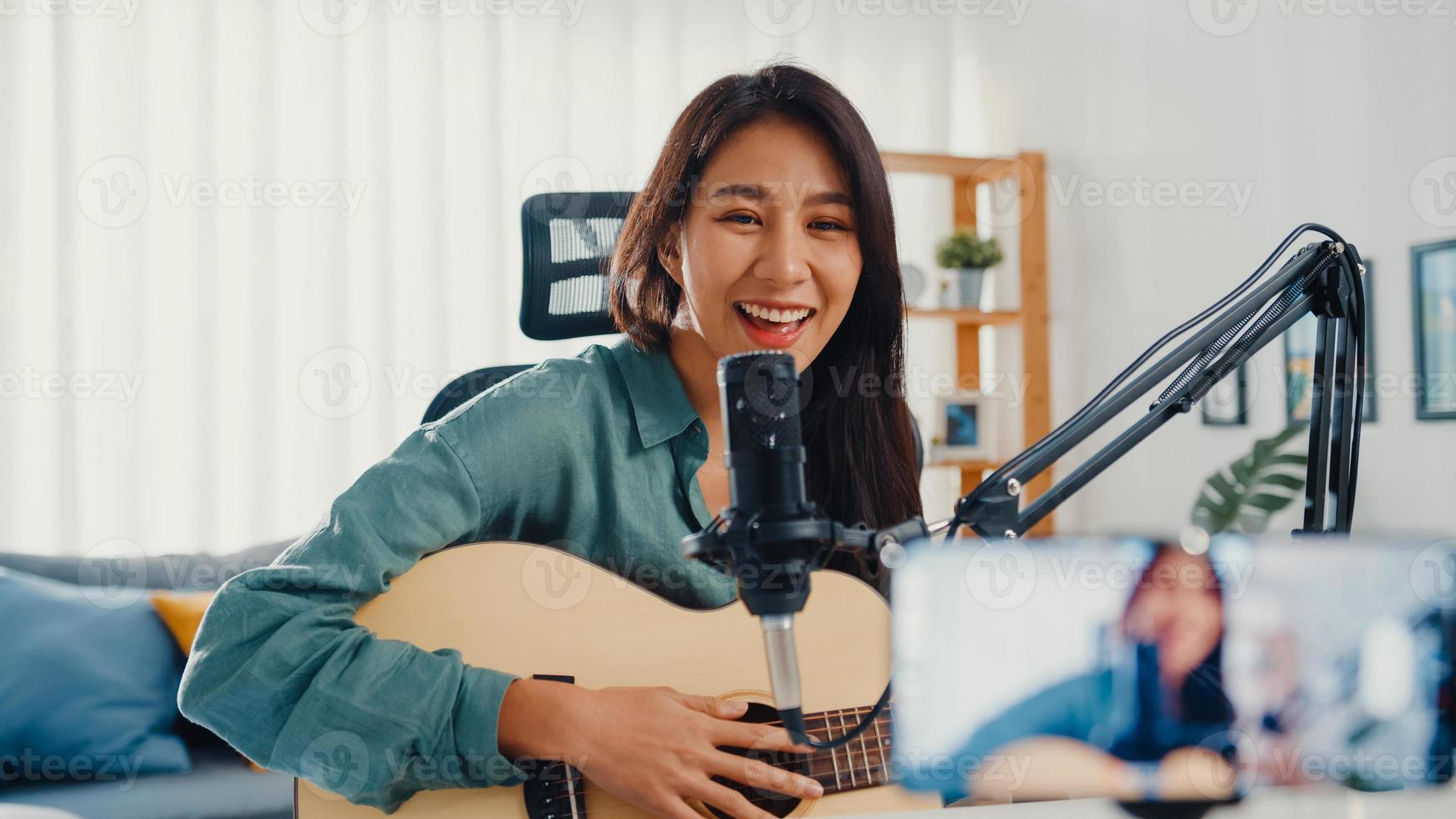 Teenage Asia girl influencer play guitar music use microphone record with smartphone for online audience listen at home. Female podcaster make audio podcast from her home studio, Stay at home concept. photo