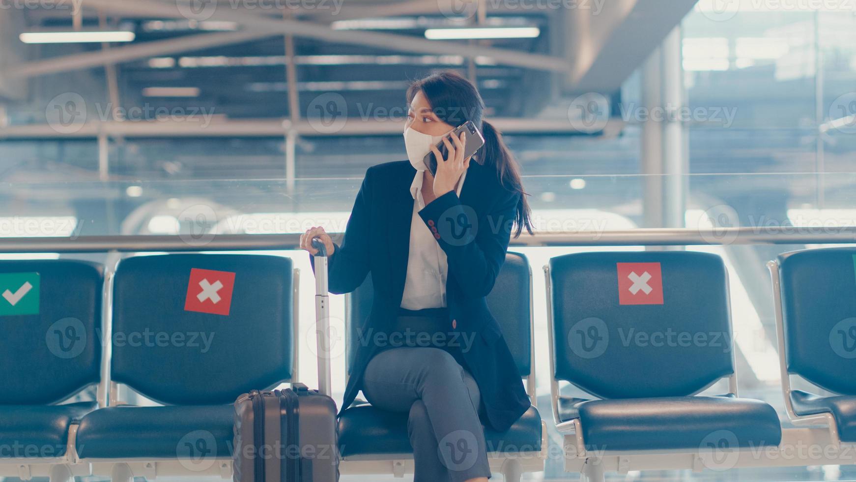 Smart asian business girl wear suit sitting with suitcase in bench call smart phone talk with partner wait for flight at airport. Business travel commuter in covid pandemic, Business travel concept. photo