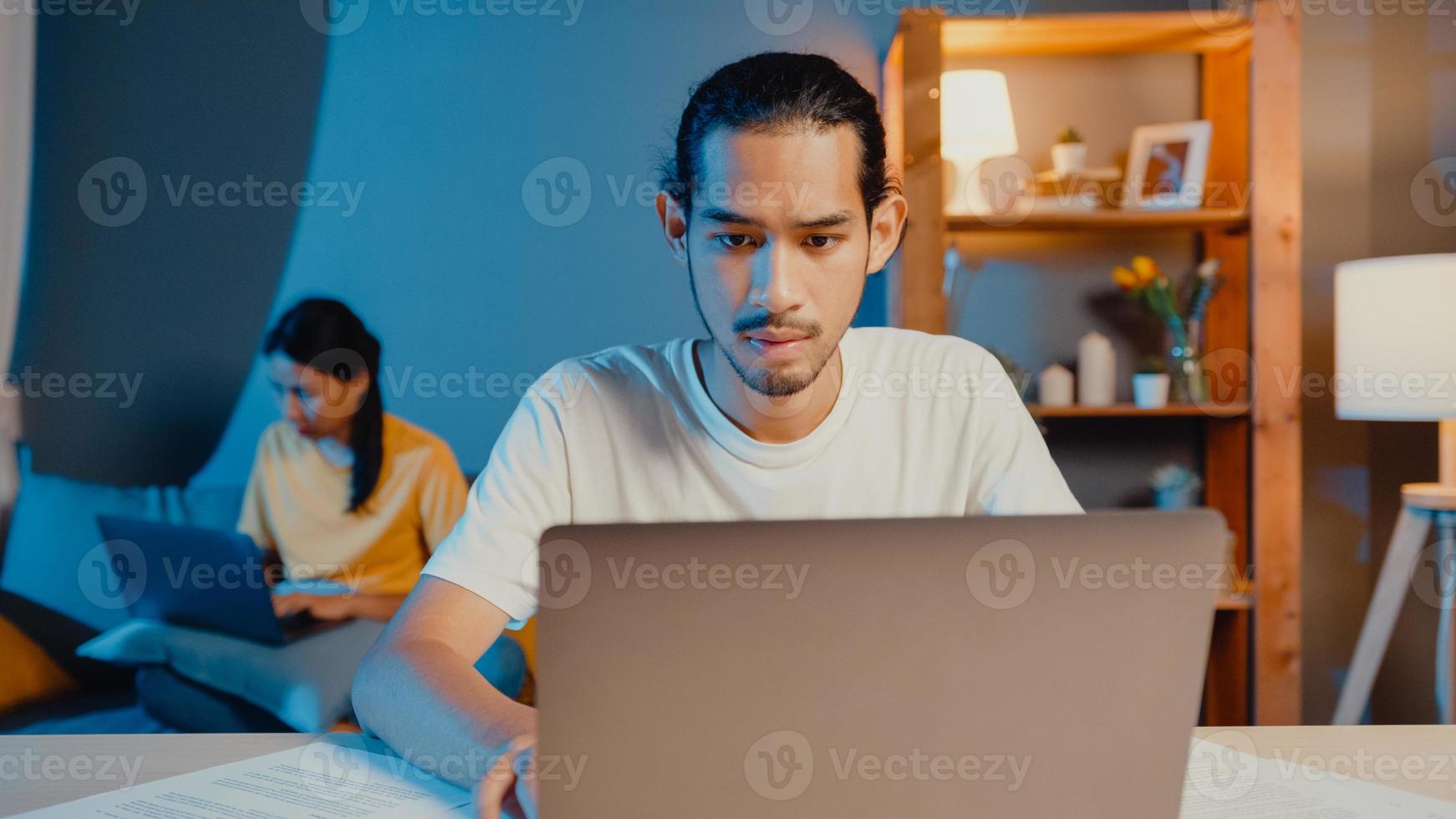 freelance asia pareja hombre y mujer en casual se sienten seriamente centrados en el trabajo computadora portátil separada por la noche, hasband sentarse frente a la mesa esposa sentarse detrás en el sofá de la casa. trabajar desde el concepto de hogar. foto