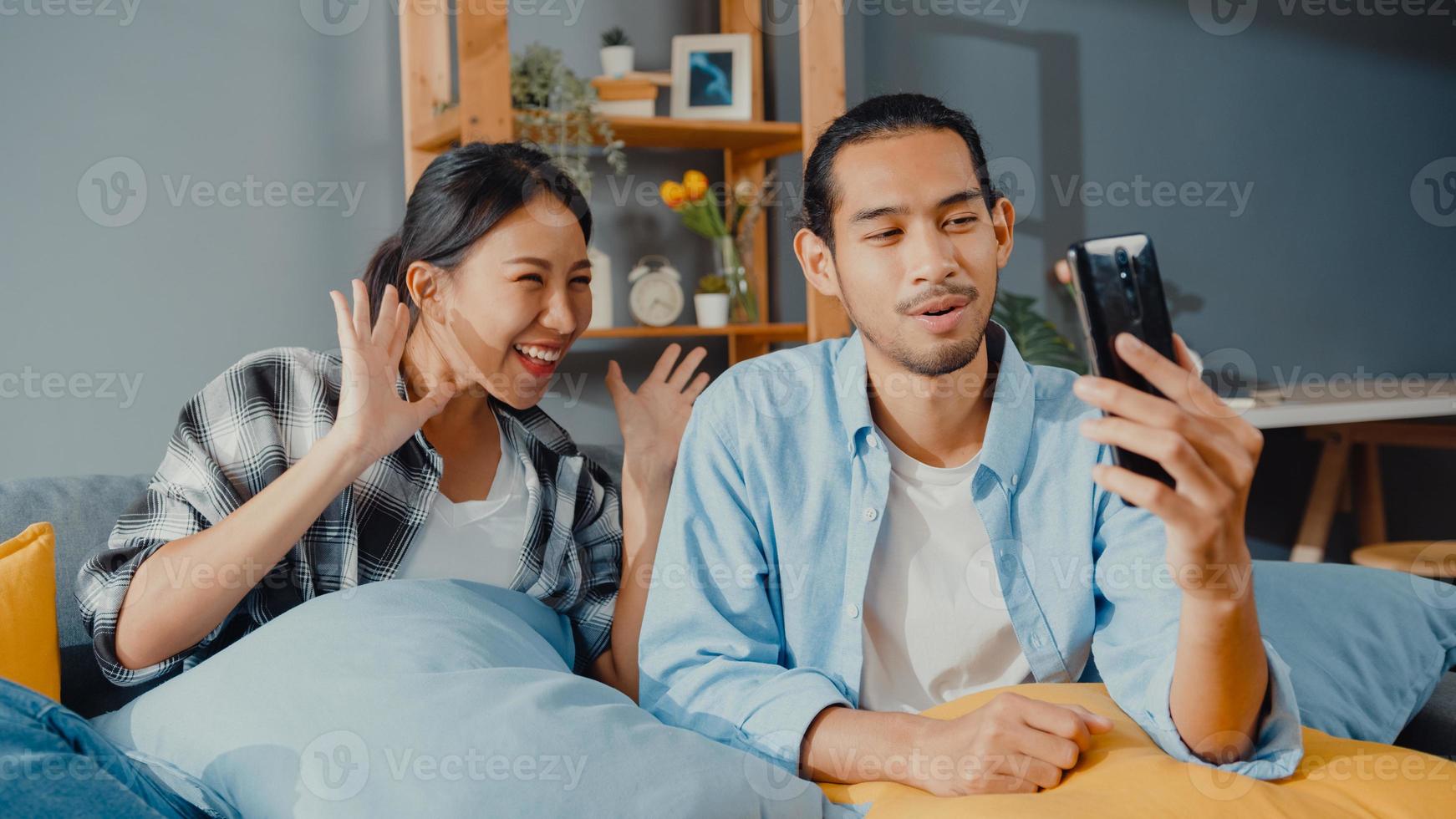 Happy young asian couple man and woman sit couch use smartphone facetime video call with friends and family in living room at home. Stay at home quarantine, Social distancing, Young married concept. photo