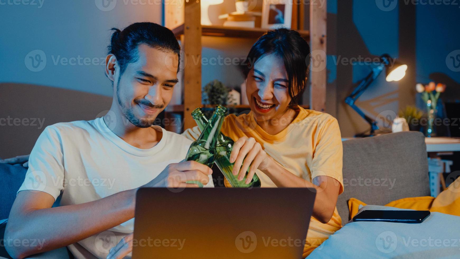 Feliz pareja asiática joven disfruta de un evento de fiesta nocturna en línea, sentarse en el sofá, usar una videollamada de computadora portátil con amigos, tostar, beber cerveza a través de una videollamada en línea en la sala de estar en casa, concepto de distanciamiento social. foto