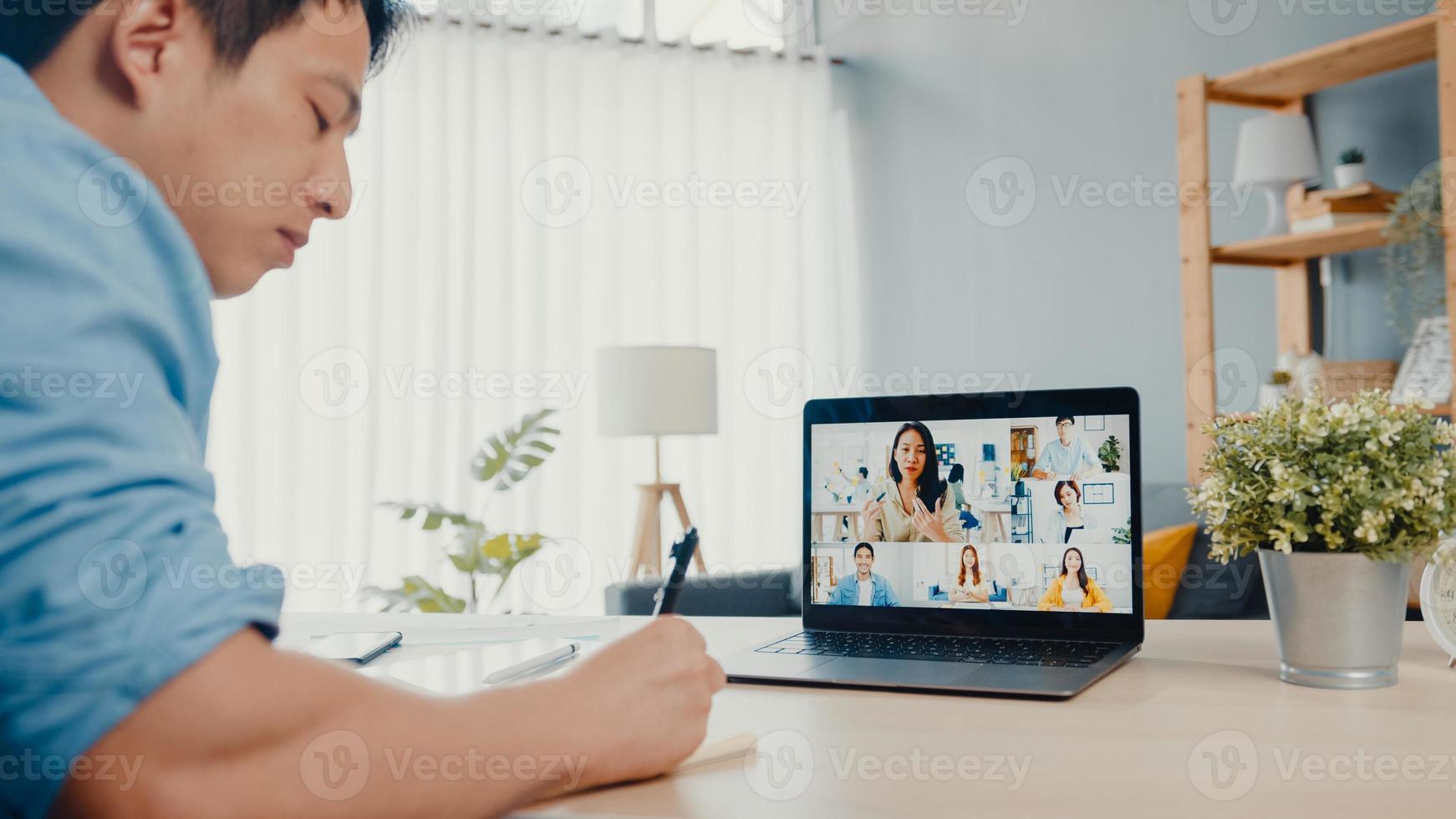 Young Asia businessman using laptop talk to colleagues about plan in video call meeting while work from home at living room. Self-isolation, social distancing, quarantine for corona virus prevention. photo