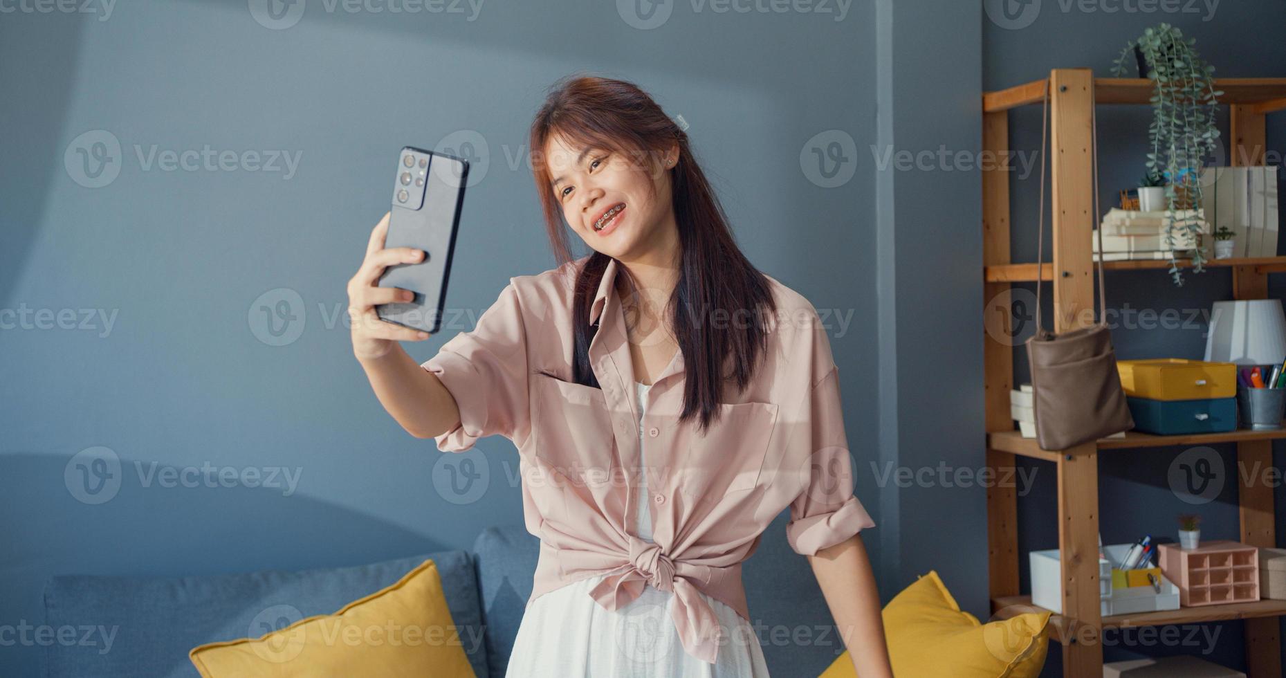 Happy carefree young Asia girl use smartphone video call enjoy talk with college friend in living room at home. Social distance coronavirus pandemic concept. Freedom and active lifestyle concept. photo