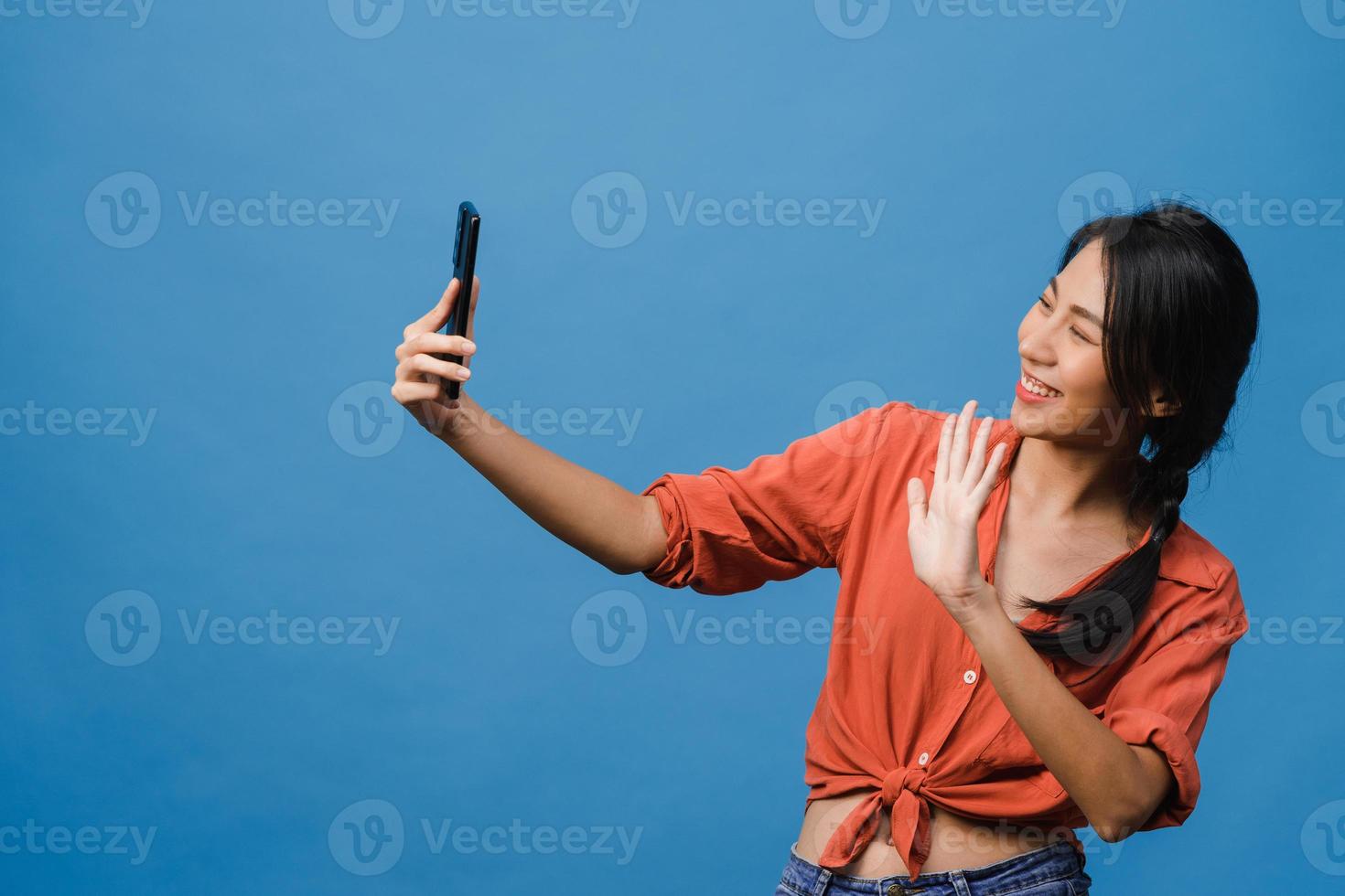 Smiling adorable Asian female making selfie photo on smart phone with positive expression in casual clothing and stand isolated on blue background. Happy adorable glad woman rejoices success.