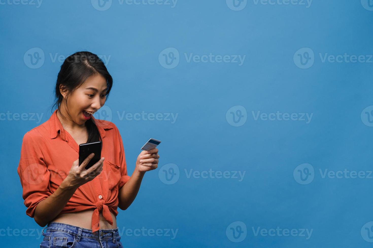 Jovencita asiática usando teléfono y tarjeta bancaria de crédito con expresión positiva, sonríe ampliamente, vestida con ropa informal y está aislada sobre fondo azul. feliz adorable mujer alegre se regocija con el éxito. foto