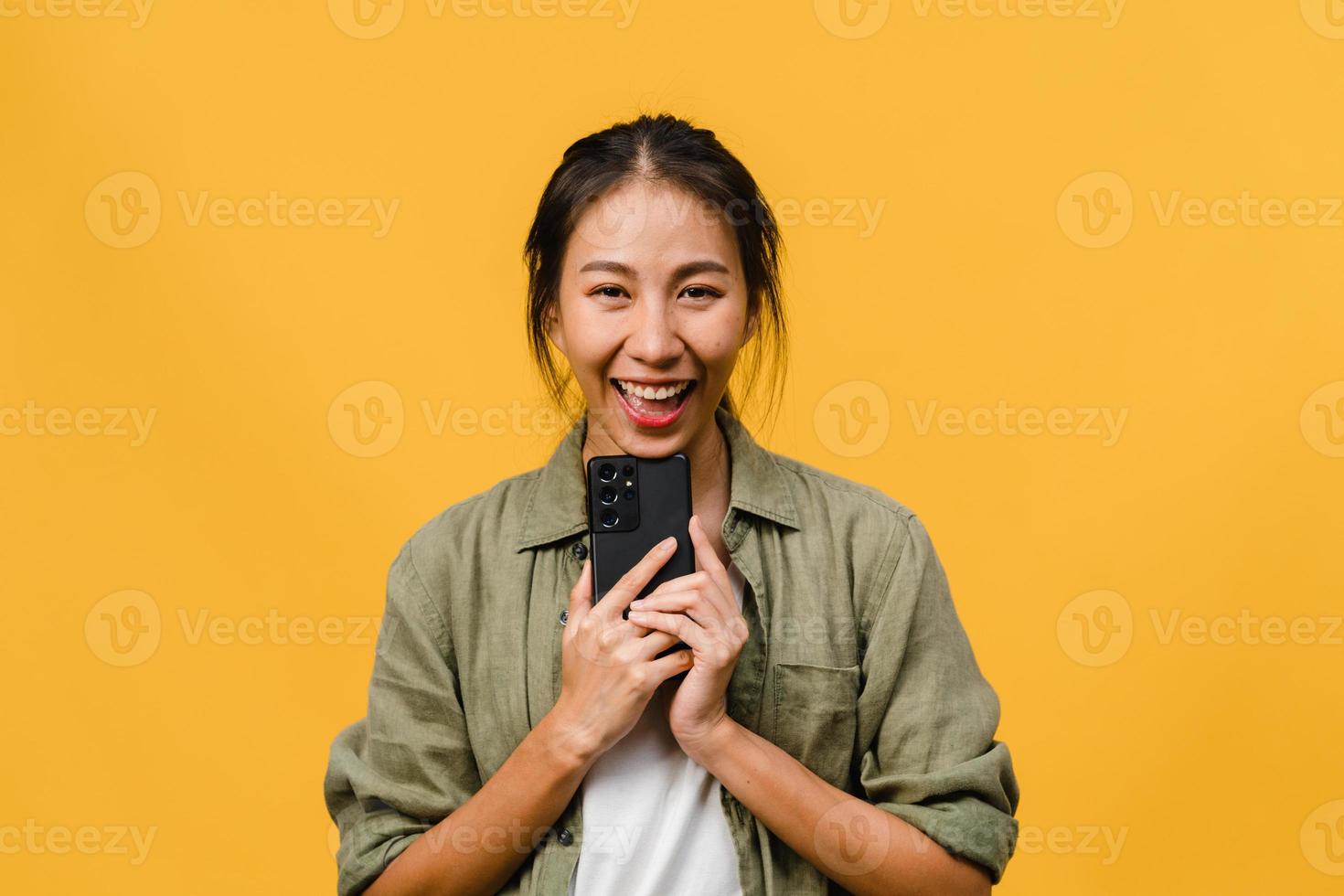 Surprised young Asia lady using mobile phone with positive expression, smile broadly, dressed in casual clothing and looking at camera on yellow background. Happy adorable glad woman rejoices success. photo