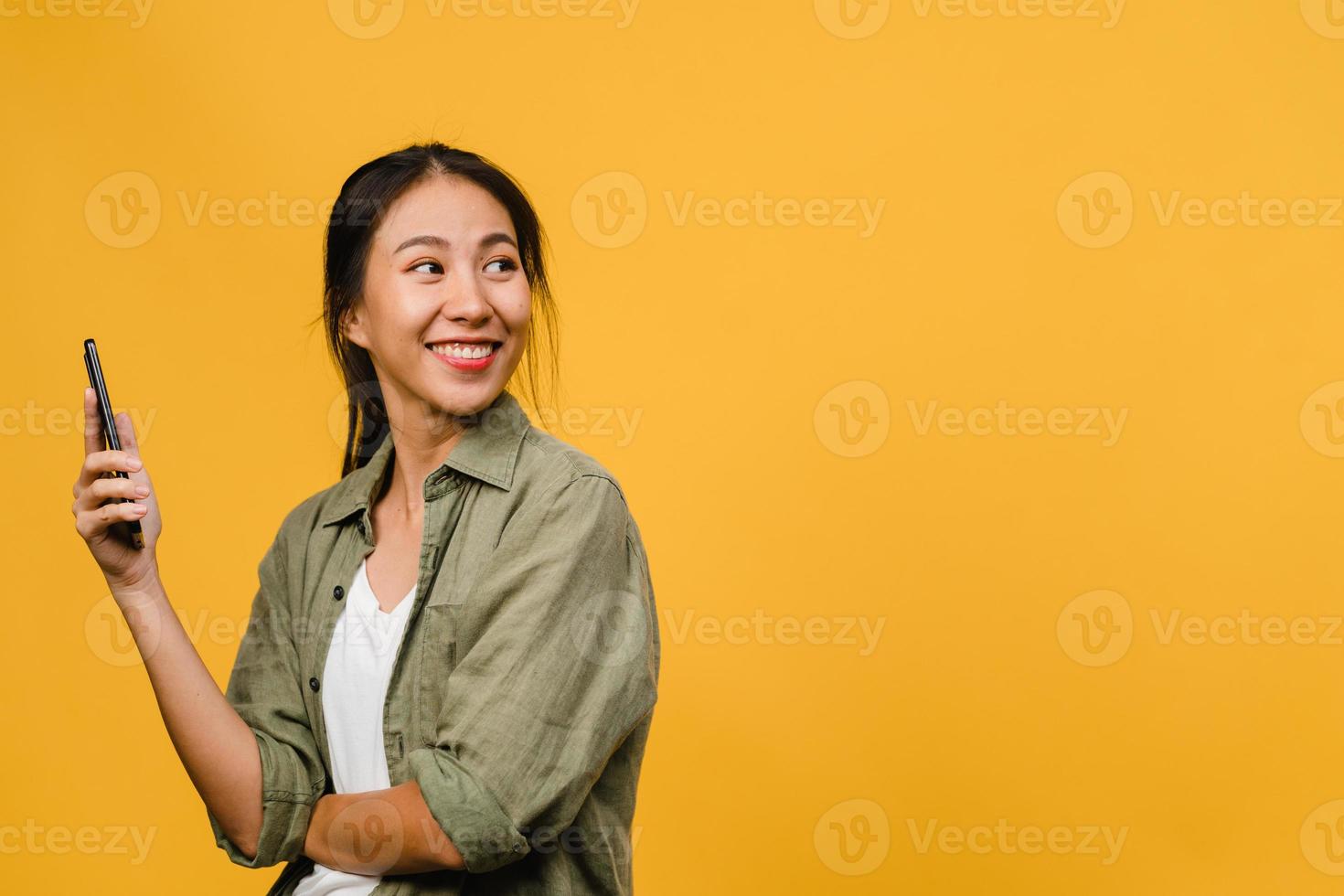 Jovencita asiática que usa el teléfono con expresión positiva, sonríe ampliamente, vestida con ropa casual sintiendo felicidad y parada aislada sobre fondo amarillo. feliz adorable mujer alegre se regocija con el éxito. foto