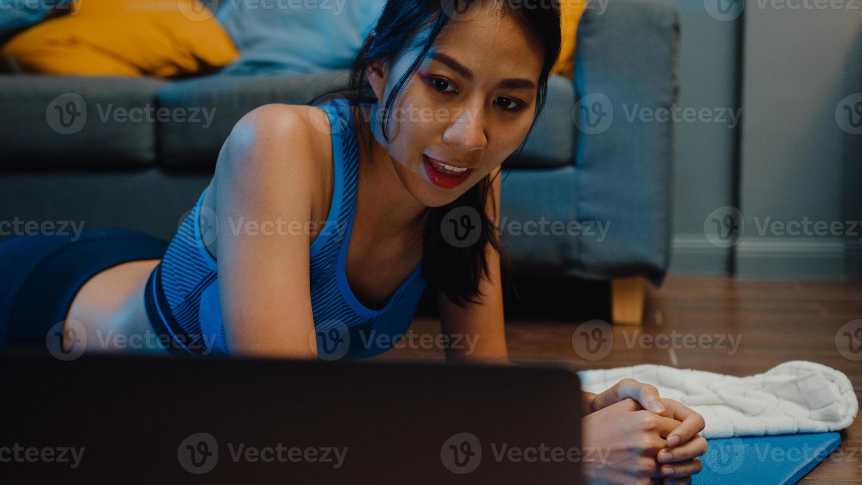 Young Asia lady in sportswear exercises doing work out and using laptop to watch yoga video tutorial at home night. Distant training with personal trainer, social distance, online education concept. photo