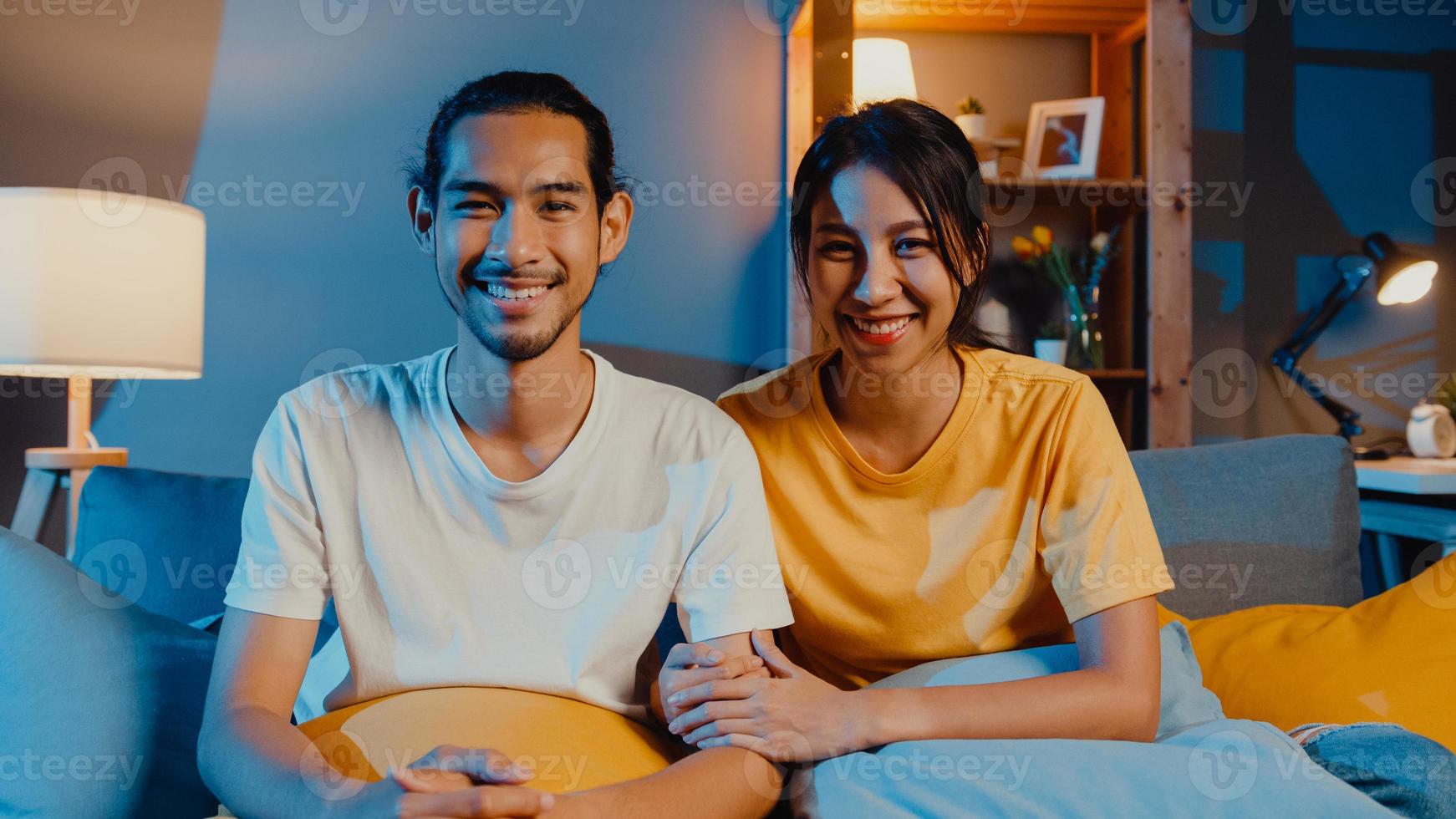 Feliz pareja de jóvenes asiáticos hombre y mujer mirando a la cámara sonríen y alegres en la videollamada en línea por la noche en la sala de estar en casa, quedarse en casa en cuarentena, vida matrimonial, concepto de distanciamiento social. foto