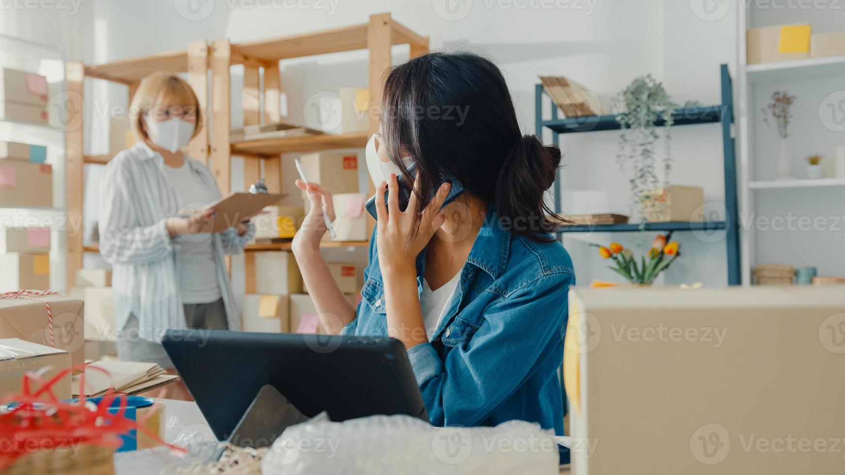 Jóvenes empresarias asiáticas usan mascarilla usando una llamada de teléfono móvil, reciben una orden de compra y verifican el producto en el trabajo de stock en la oficina en casa. propietario de una pequeña empresa, concepto independiente de entrega de mercado en línea. foto