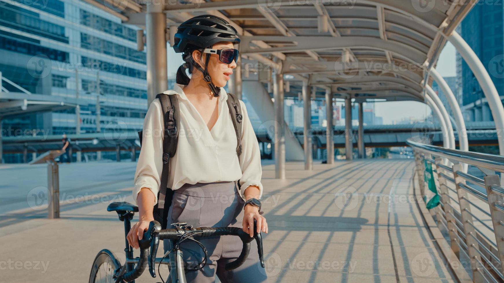 mujer asiática mujer de negocios usar gafas de sol ir a trabajar en la oficina caminar y sonreír mirar a su alrededor sostenga un soporte para bicicletas alrededor del edificio en una calle de la ciudad. viaje en bicicleta, viaje en bicicleta, concepto de viajero de negocios. foto