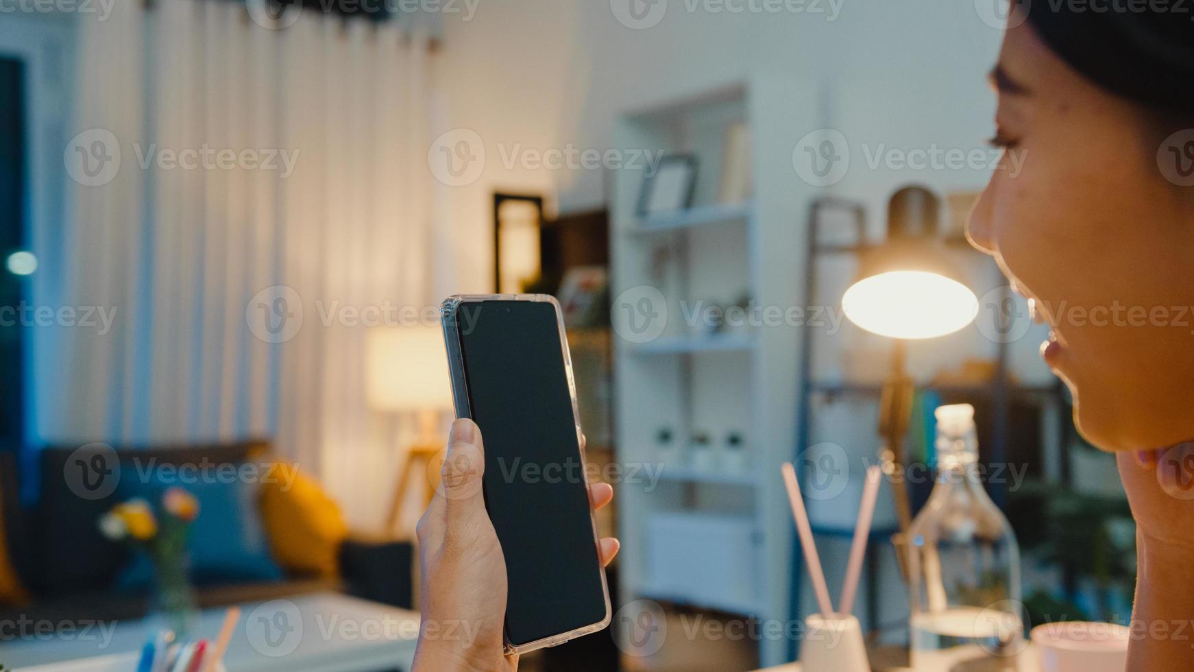 La joven asiática usa un teléfono inteligente con pantalla negra en blanco para mostrar texto publicitario mientras trabaja de manera inteligente desde su casa en la sala de estar por la noche. tecnología chroma key, concepto de diseño de marketing. foto