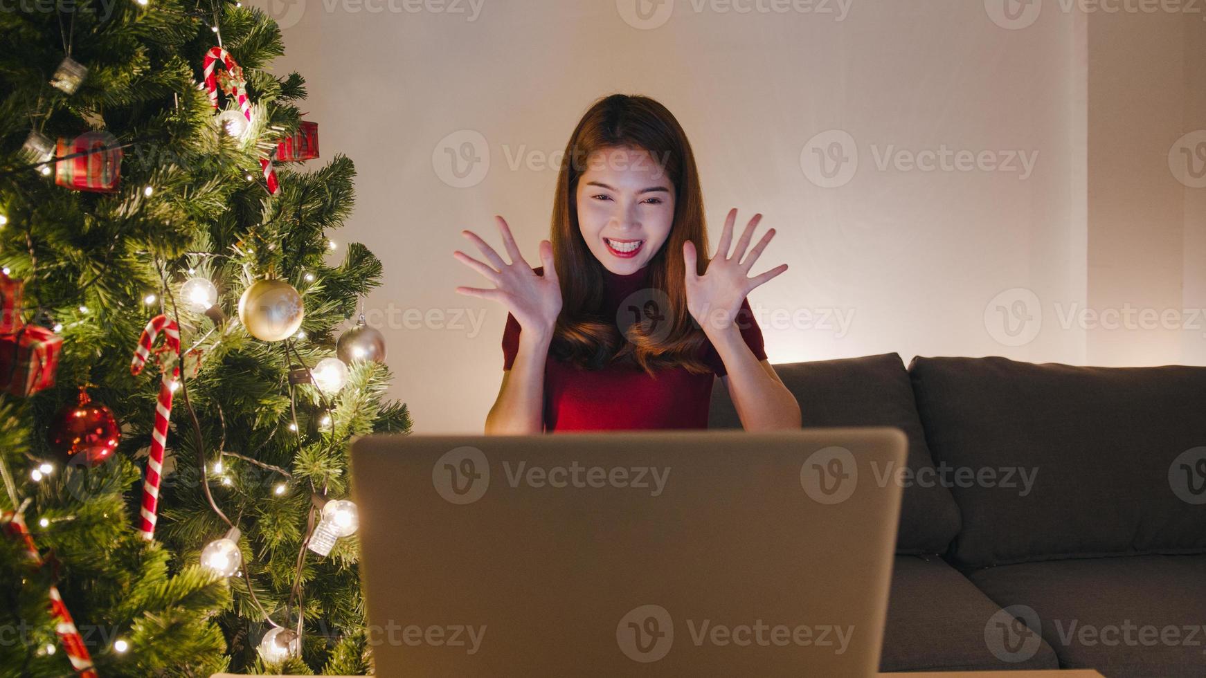 Mujer joven de Asia con videollamada portátil hablando con pareja con caja de regalo de Navidad, árbol de Navidad decorado con adornos en la sala de estar en casa. noche de navidad y fiesta de año nuevo. foto