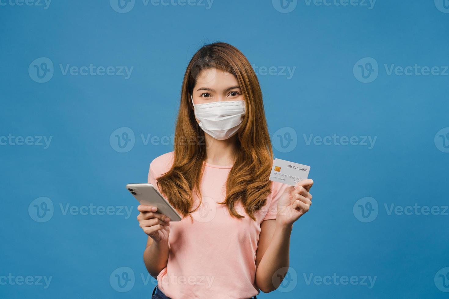 joven asiática con mascarilla médica usando teléfono y tarjeta bancaria de crédito con expresión positiva, sonríe ampliamente, vestida con ropa casual y de pie aislado sobre fondo azul. foto