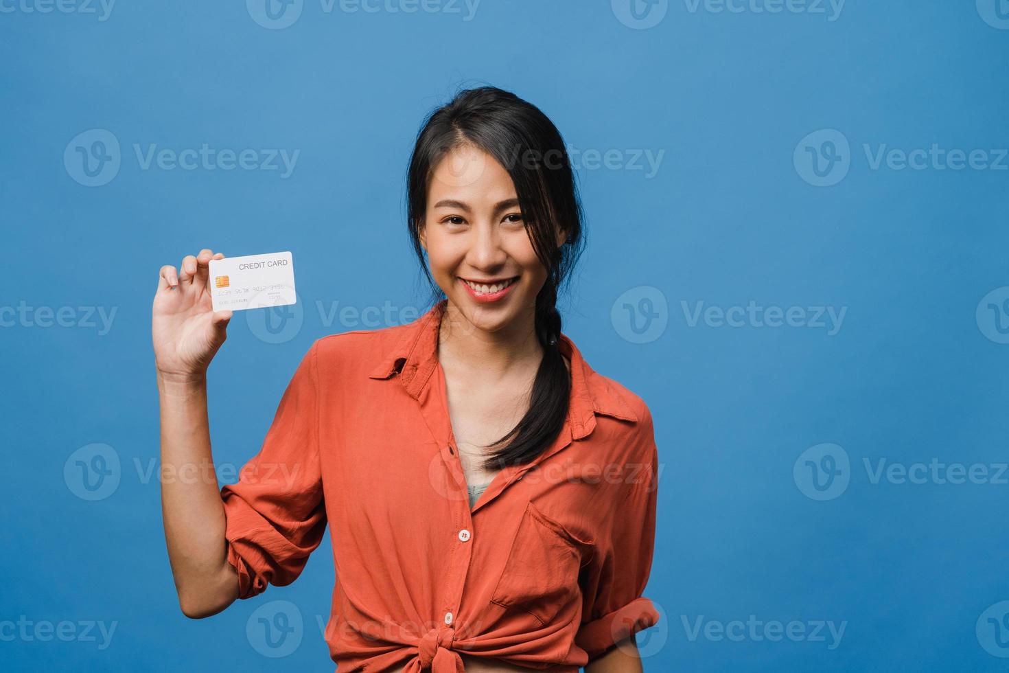 Joven mujer asiática muestra una tarjeta bancaria de crédito con expresión positiva, sonríe ampliamente, vestida con ropa informal sintiendo felicidad y parada aislada sobre fondo azul. concepto de expresión facial. foto