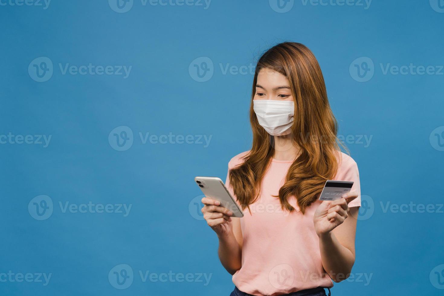 joven asiática con mascarilla médica usando teléfono y tarjeta bancaria de crédito con expresión positiva, sonríe ampliamente, vestida con ropa casual y de pie aislado sobre fondo azul. foto