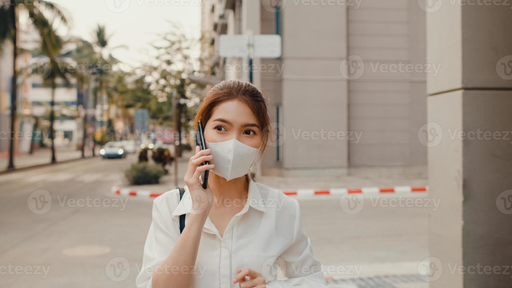 Successful young Asia businesswoman in fashion office clothes wear medical face mask talking via mobile phone while walking alone outdoor in urban modern city in morning. Business on the go concept. photo