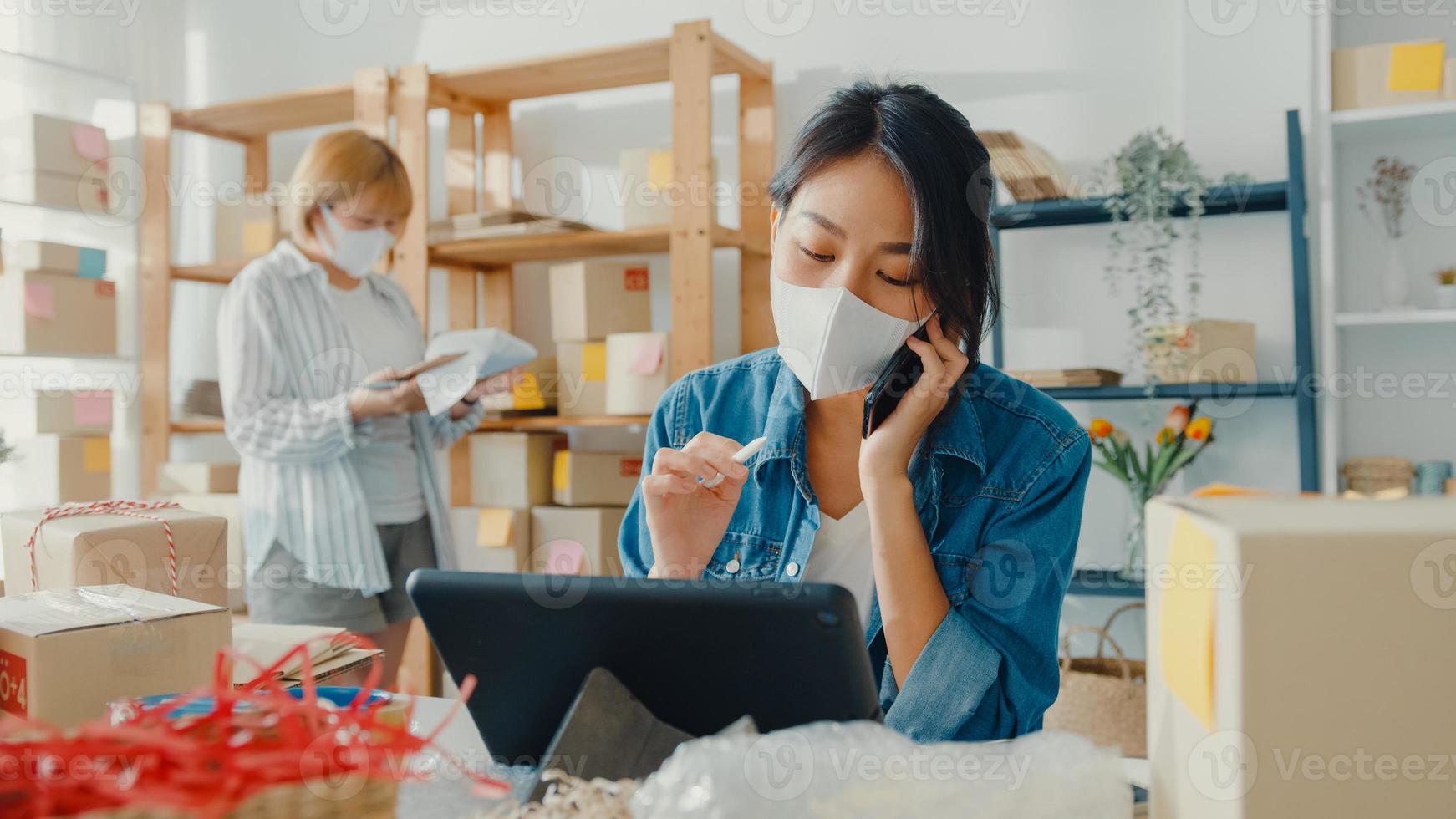 Jóvenes empresarias asiáticas usan mascarilla usando una llamada de teléfono móvil, reciben una orden de compra y verifican el producto en el trabajo de stock en la oficina en casa. propietario de una pequeña empresa, concepto independiente de entrega de mercado en línea. foto