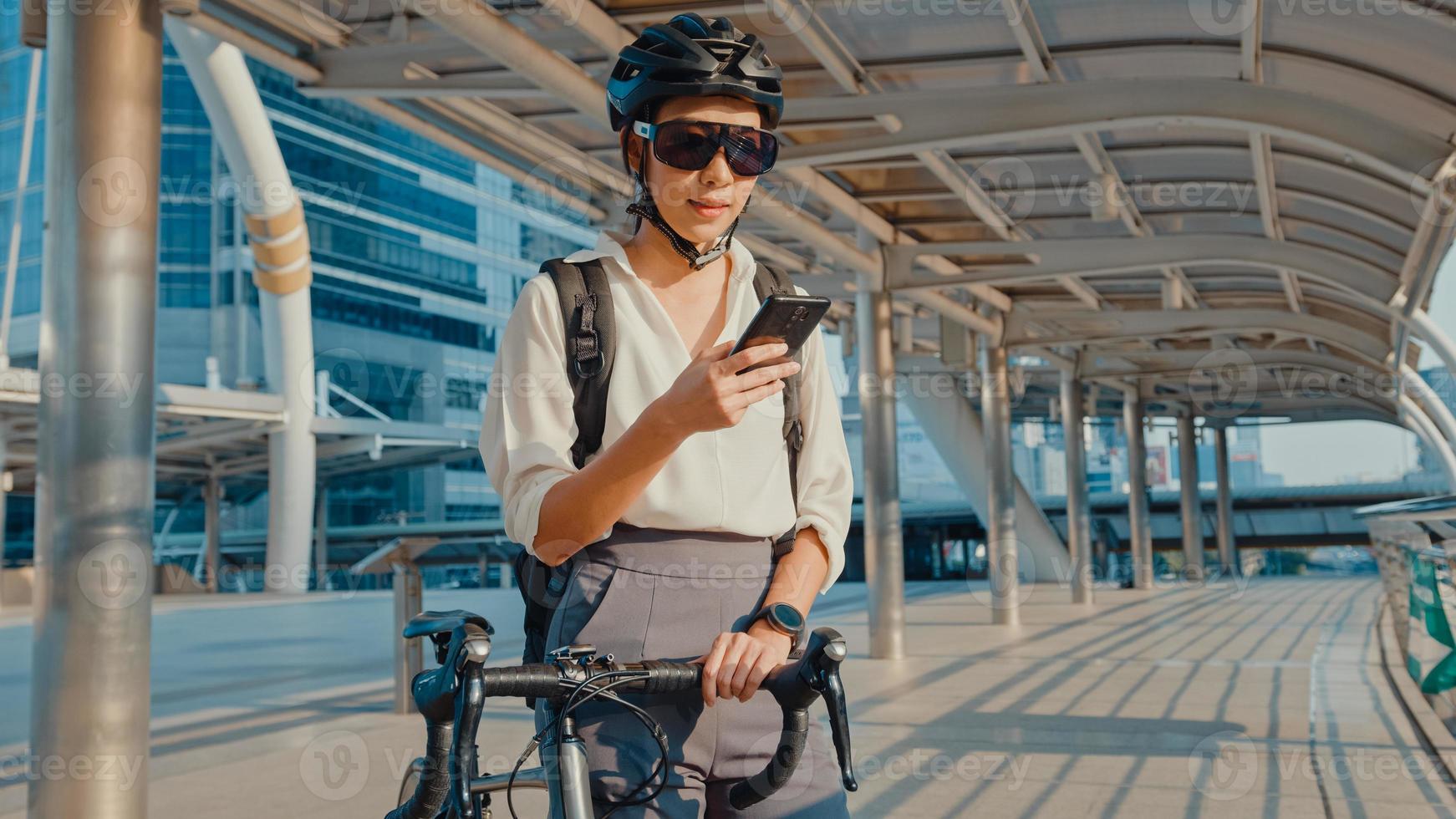 feliz empresaria asiática con mochila utiliza teléfonos inteligentes en la ciudad de pie en la calle con bicicleta ir a trabajar en la oficina. chica deportiva usa su teléfono para trabajar. viaje diario al trabajo, viajero de negocios en la ciudad. foto