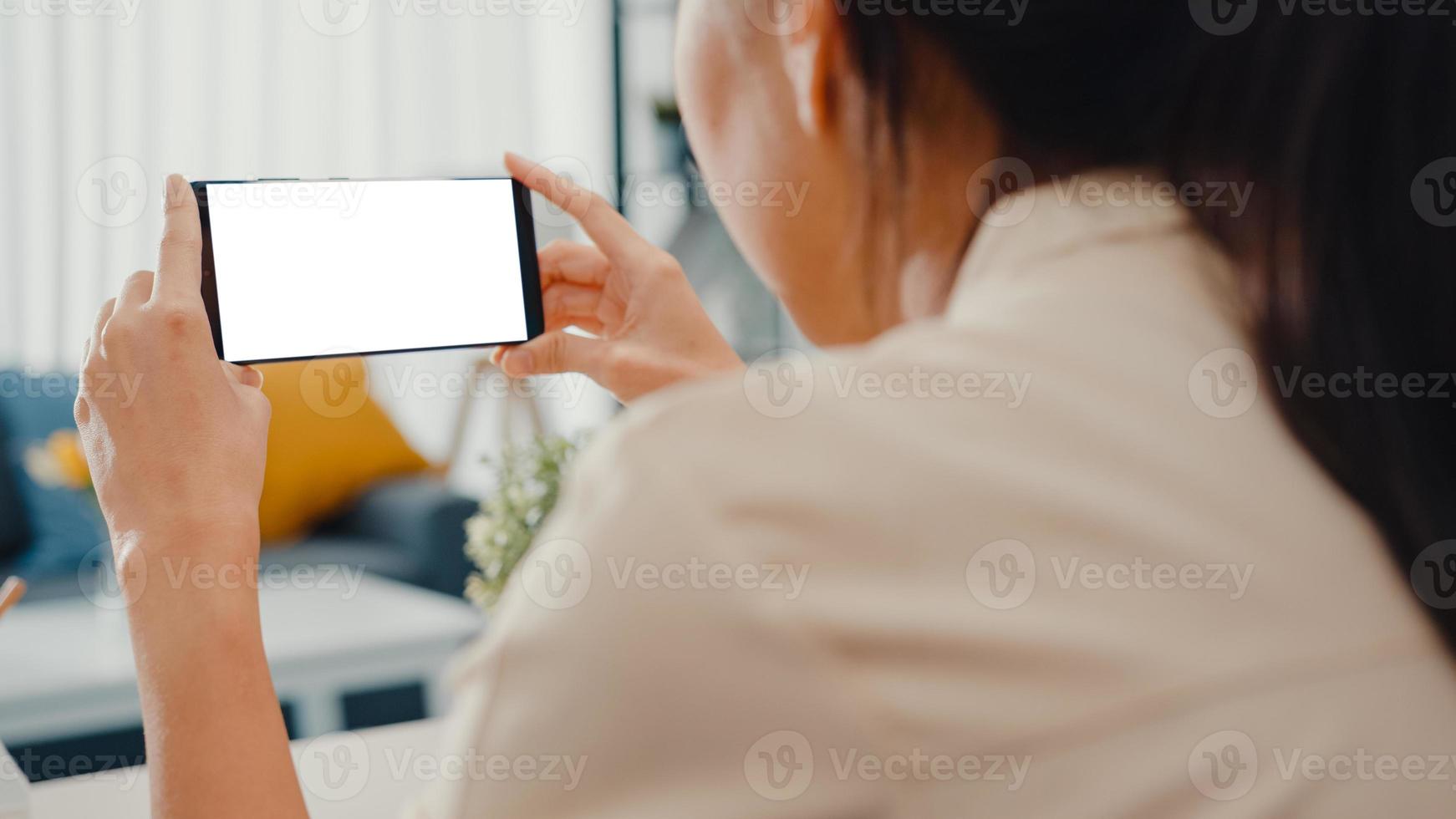 Jovencita asiática usa un teléfono inteligente con una pantalla en blanco en blanco simulado para mostrar texto publicitario mientras trabaja de manera inteligente desde casa en la sala de estar. tecnología chroma key, concepto de diseño de marketing. foto