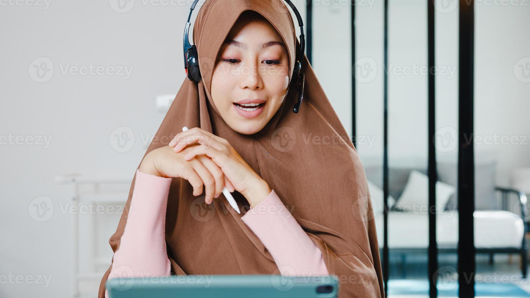 Asia muslim lady wear headphone using digital tablet talk to colleagues about sale report in conference video call while working from home at kitchen. Social distancing, quarantine for corona virus. photo