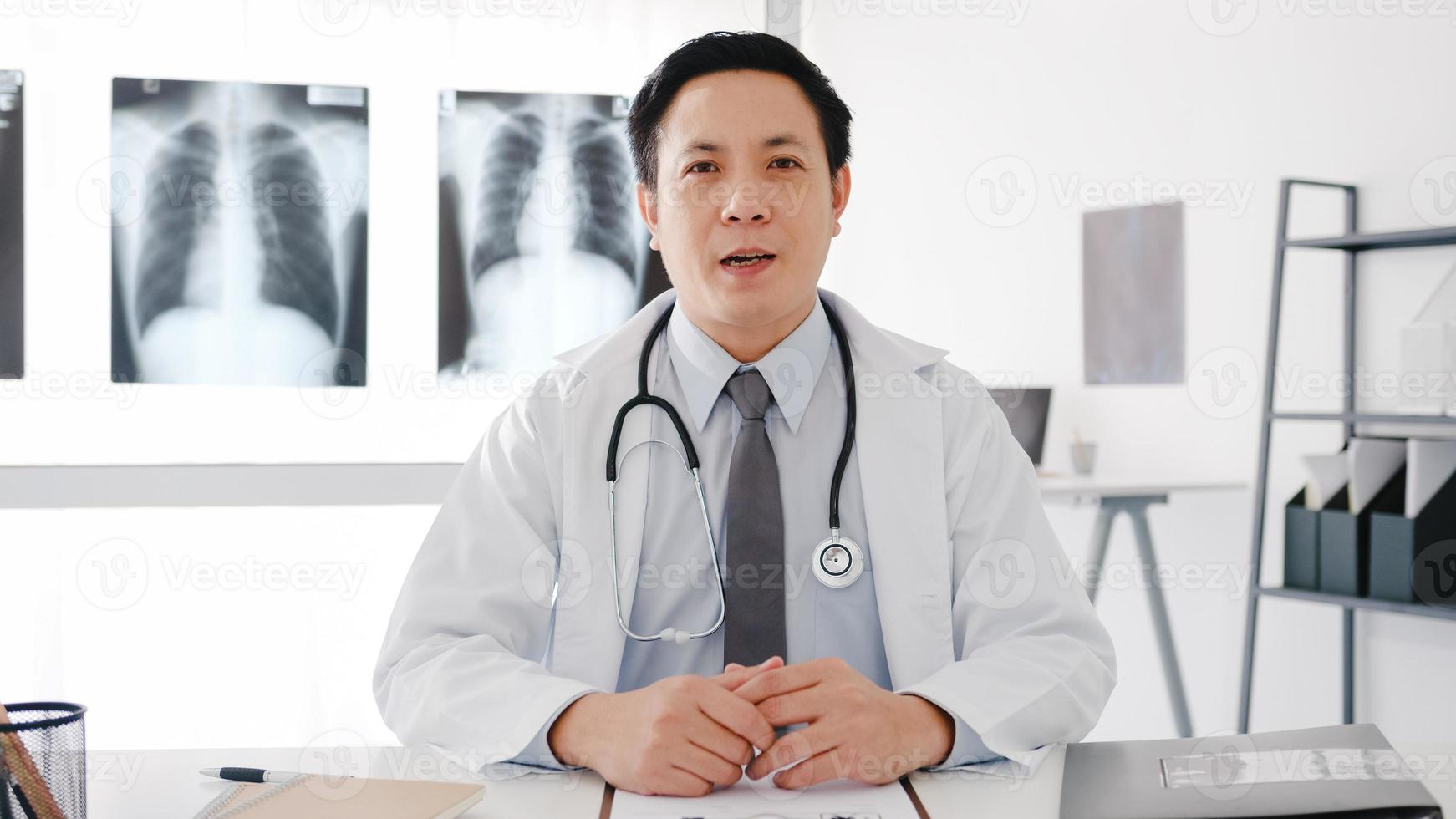 Young Asia male doctor in white medical uniform with stethoscope using computer laptop talk video conference call with patient, looking at camera in health hospital. Consulting and therapy concept. photo