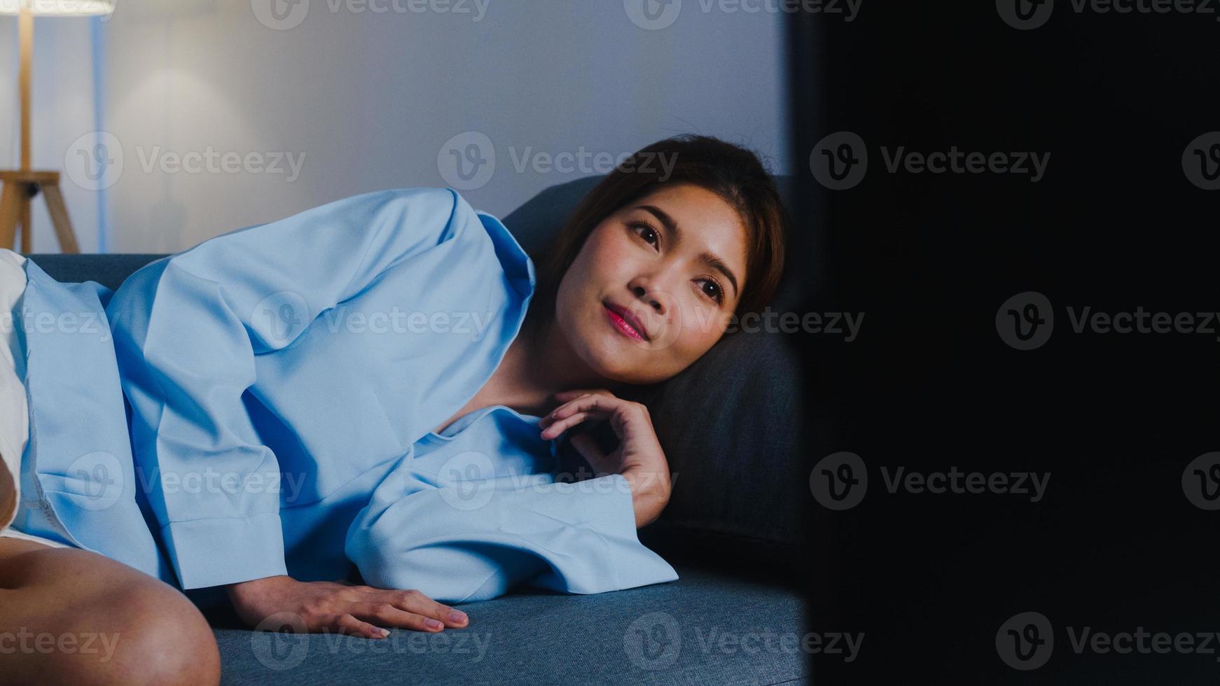 Young Asian lady watching tv feeling happy fun laugh looking television series movie while lying sofa in living room at home at night. Social distancing, quarantine for coronavirus prevention concept. photo