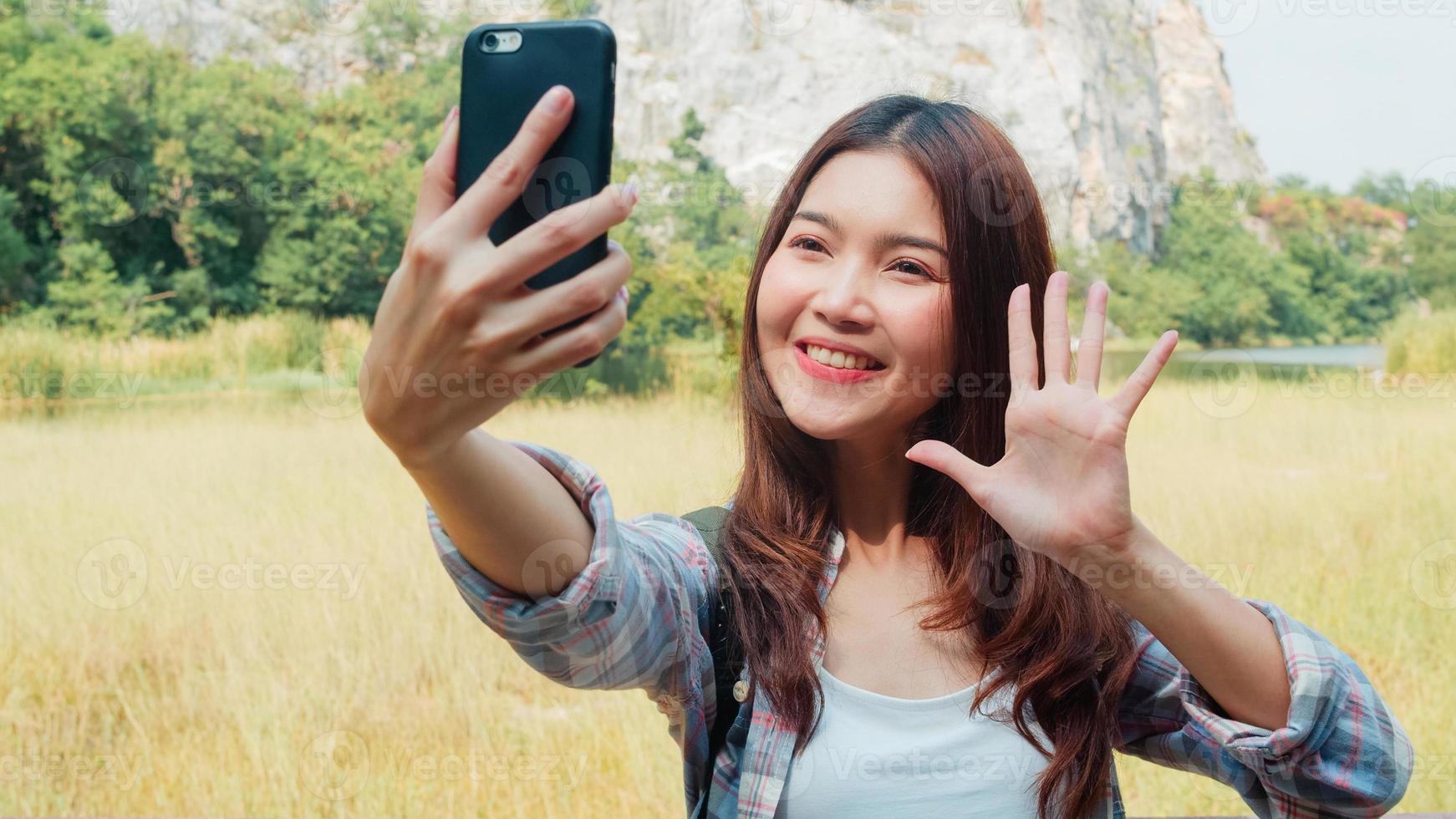 Cheerful young traveler Asian lady with backpack selfie at mountain lake. Korean girl happy using mobile phone taking selfie enjoy holidays on hiking adventure. Lifestyle travel and relax concept. photo