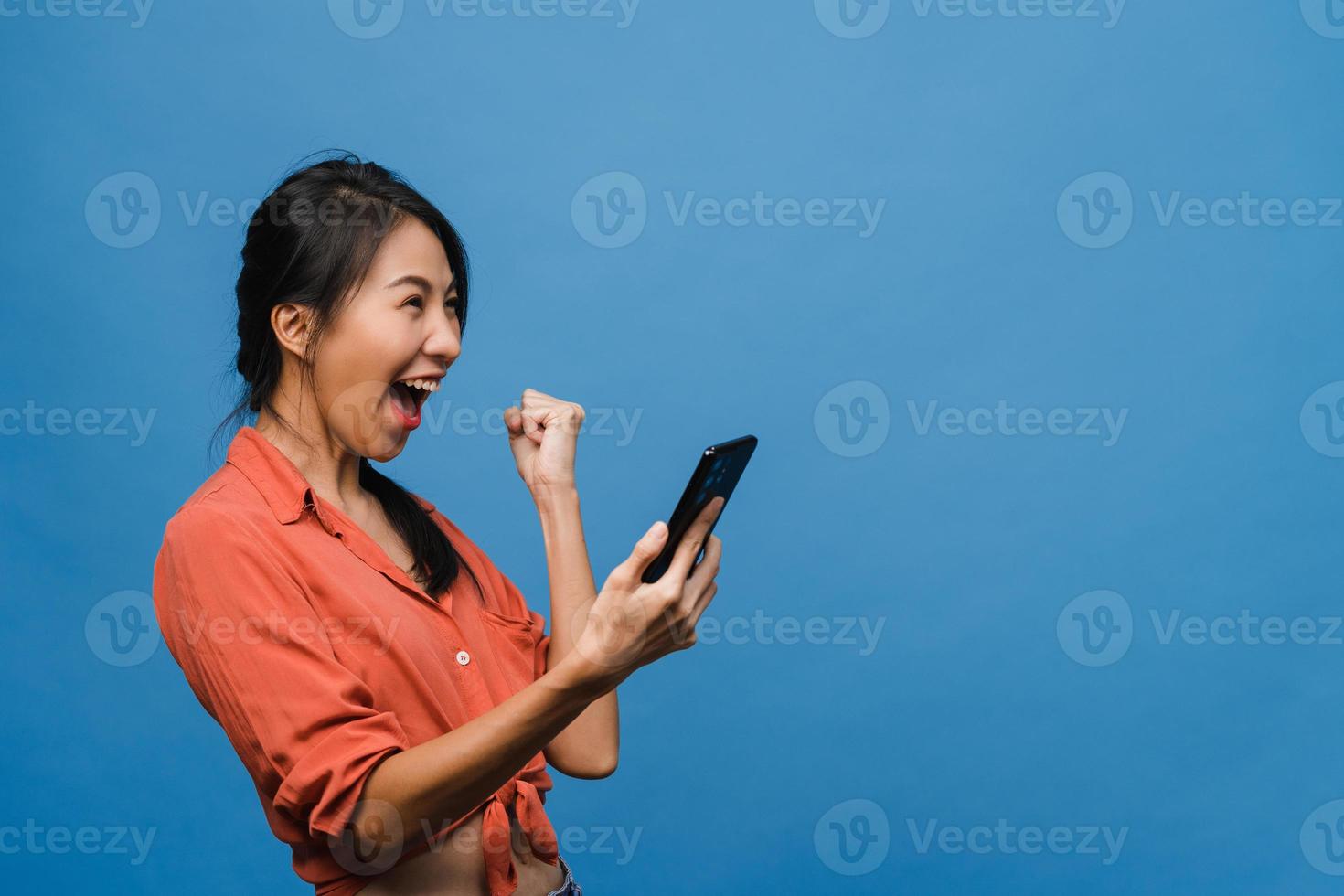 Surprised young Asia lady using mobile phone with positive expression, smiles broadly, dressed in casual clothing and stand isolated on blue background. Happy adorable glad woman rejoices success. photo