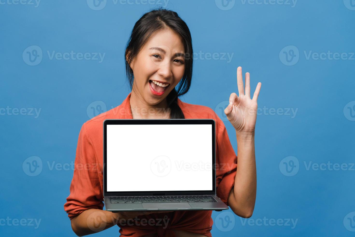 Joven asiática muestra una pantalla de computadora portátil vacía con expresión positiva, sonríe ampliamente, vestida con ropa casual sintiendo felicidad aislada sobre fondo azul. computadora con pantalla blanca en mano femenina. foto