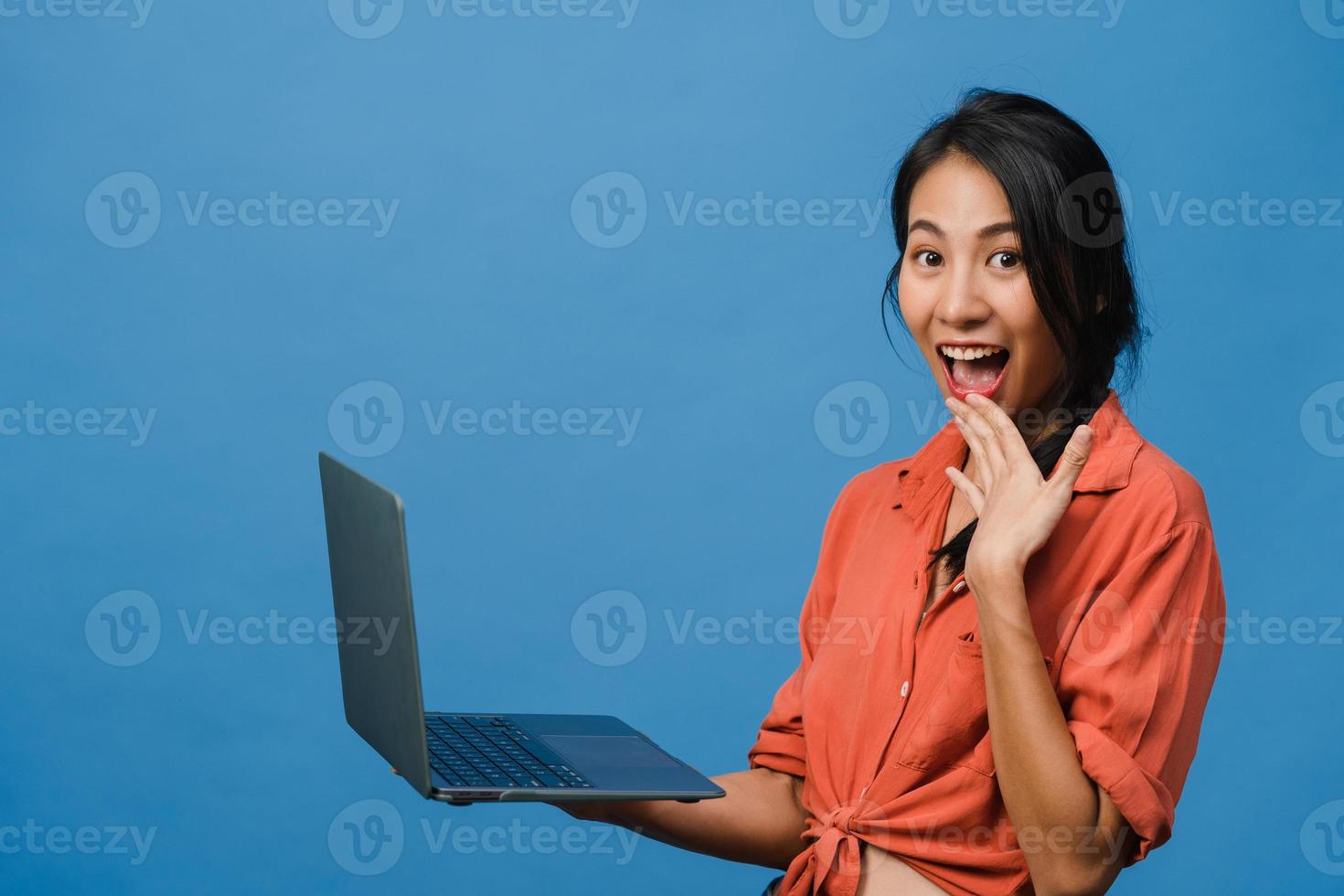 Surprised young Asia lady using laptop with positive expression, smile broadly, dressed in casual clothing and looking at camera on blue background. Happy adorable glad woman rejoices success. photo