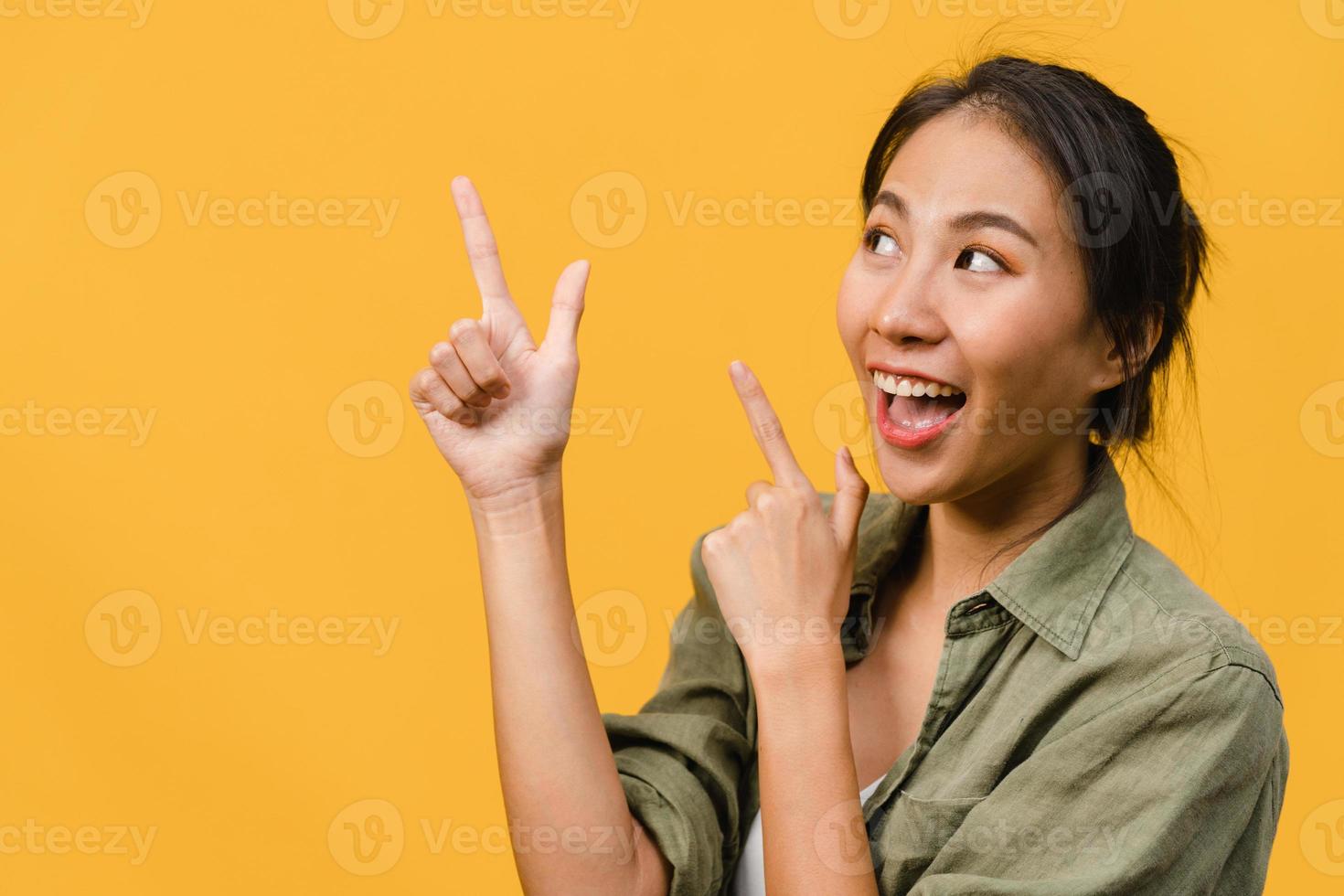 Portrait of young Asian lady smiling with cheerful expression, shows something amazing at blank space in casual clothing and standing isolated over yellow background. Facial expression concept. photo