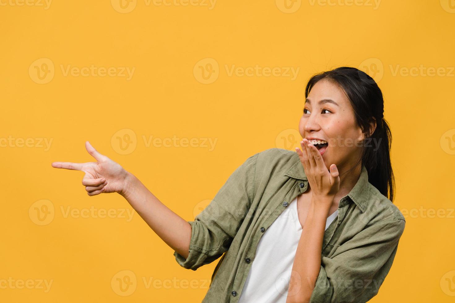 Portrait of young Asian lady smiling with cheerful expression, shows something amazing at blank space in casual clothing and standing isolated over yellow background. Facial expression concept. photo