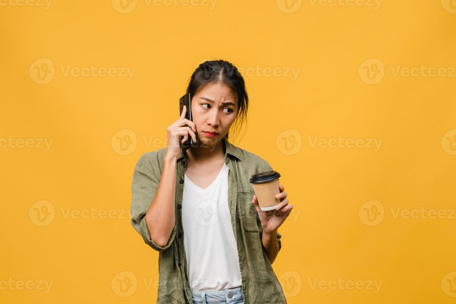 Jovencita asiática habla por teléfono y sostiene la taza de café con expresión negativa, gritos emocionados, llora enojado emocionalmente en ropa casual y se para aislado sobre fondo amarillo. concepto de expresión facial. foto