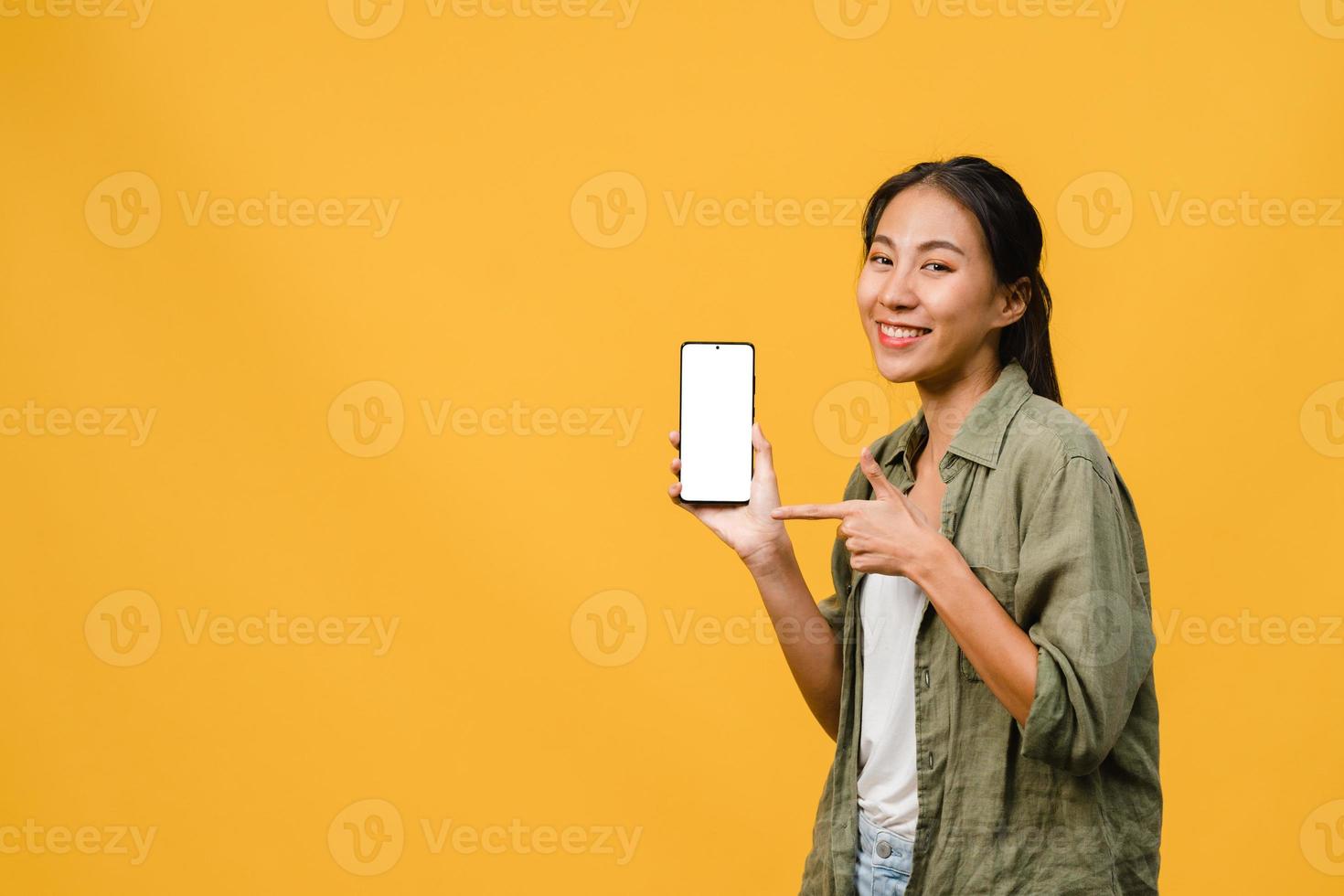 Joven asiática muestra la pantalla vacía del teléfono inteligente con expresión positiva, sonríe ampliamente, vestida con ropa casual sintiendo felicidad sobre fondo amarillo. teléfono móvil con pantalla en blanco en mano femenina. foto