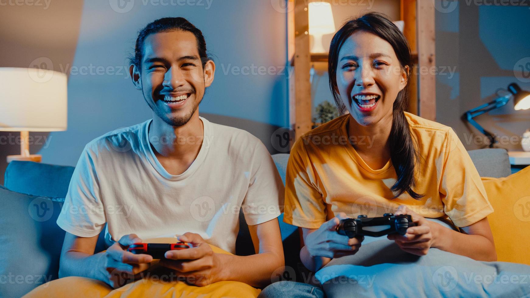 Happy asia young couple man and woman sit couch use joystick controller play video game spend fun time together in living room at night. Asian married couple family lifestyle, Stay at home concept. photo
