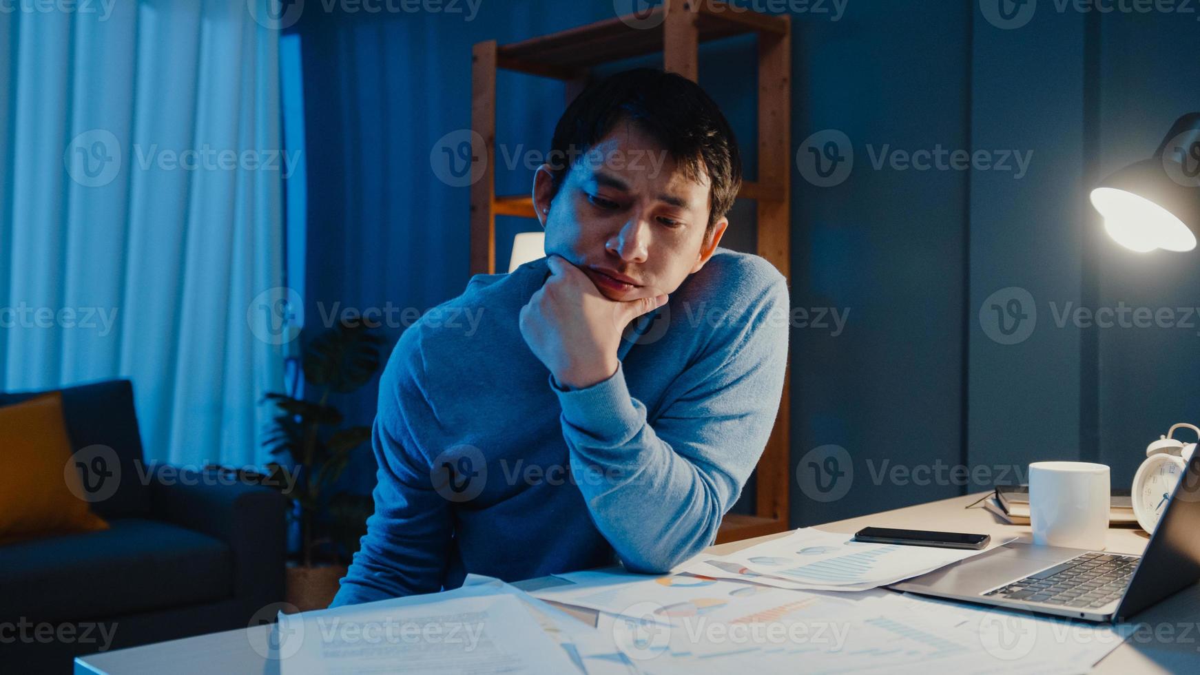 Asian freelance businessman focus work type on laptop computer busy with full of paperwork chart on desk in living room at home overtime at night, Work from home during COVID-19 pandemic concept. photo