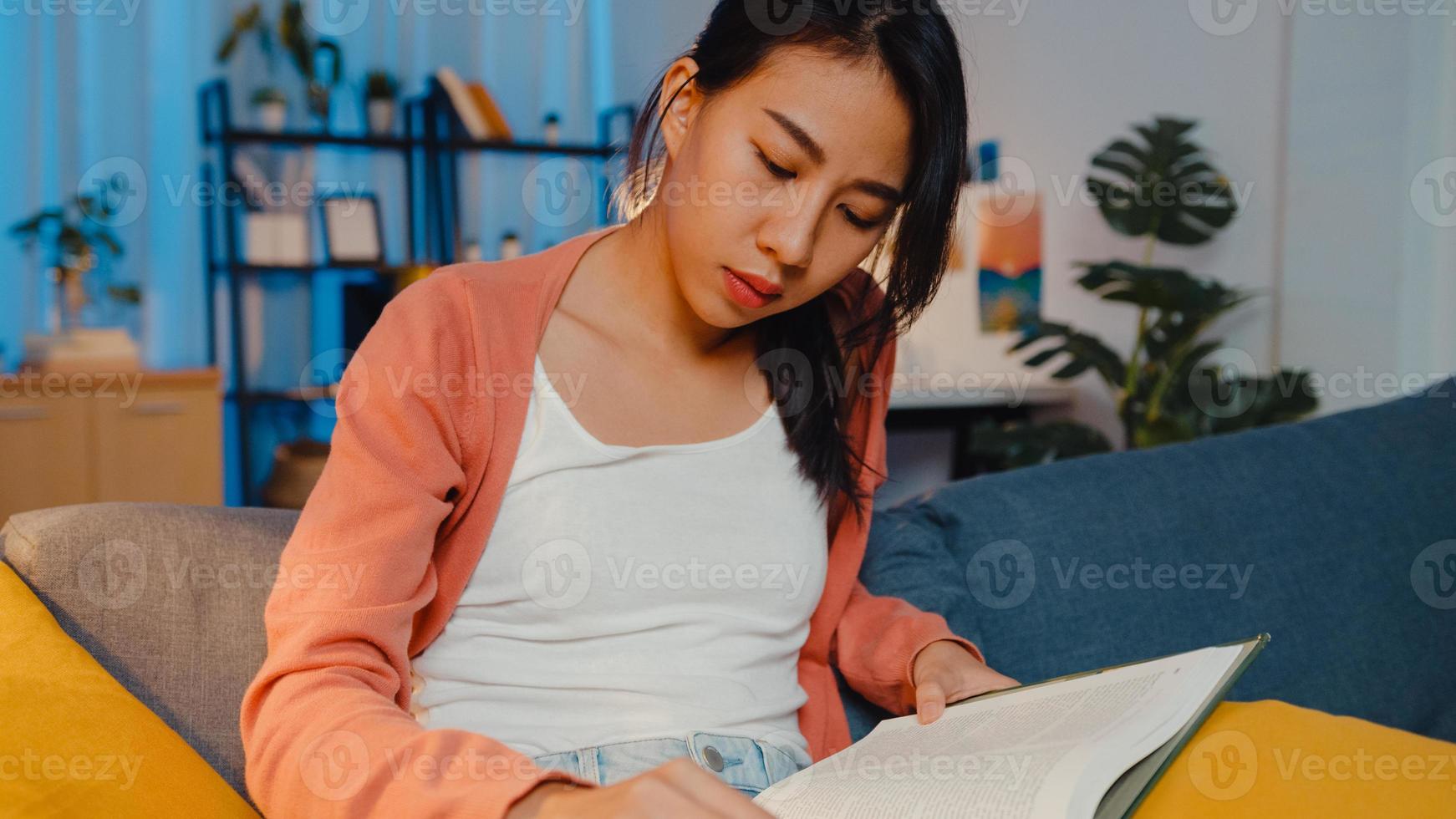 noche hermosa dama de asia lee el libro con felicidad en la tranquila sala de estar en el sofá. educación desde casa, quedarse en casa, actividad de auto cuarentena, actividad divertida para la cuarentena de covid o coronavirus. foto