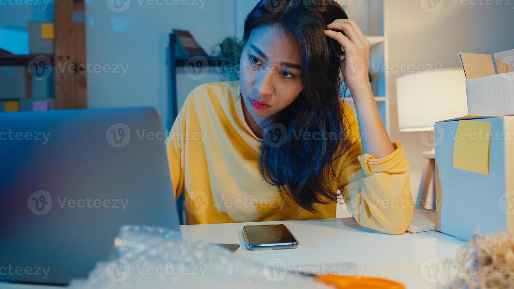 Young Asia businesswoman look around room full of product stuff and parcel box feel stress and upset with bad sell in home office at night. Small business owner, online market delivery concept. photo