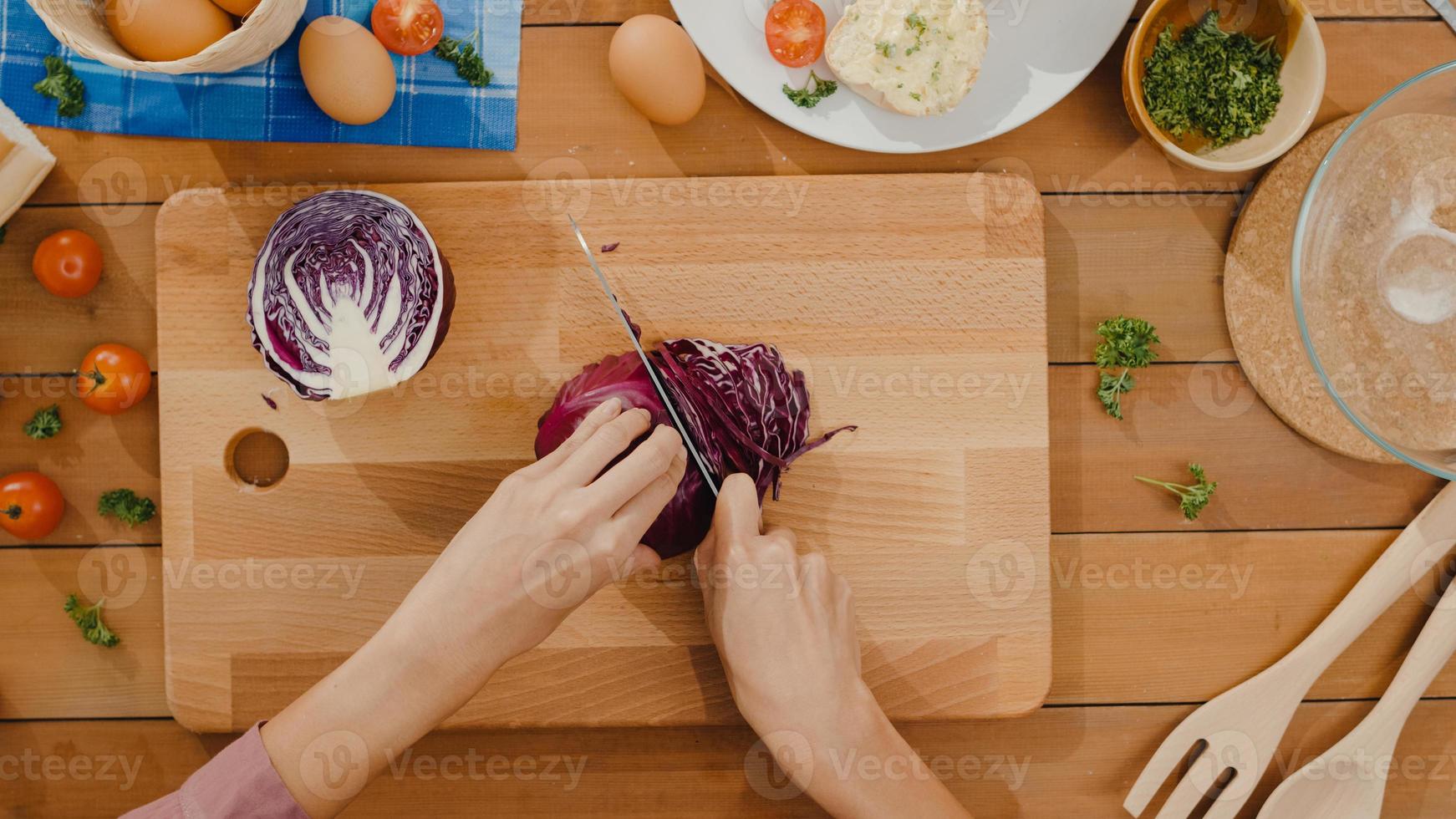 La mano del cocinero de la mujer asiática joven sostiene el cuchillo que corta el repollo rojo en la tabla de madera en la mesa de la cocina en casa. cocinar ensalada de verduras, comida sana de estilo de vida y concepto natural tradicional. tiro de la vista superior. foto