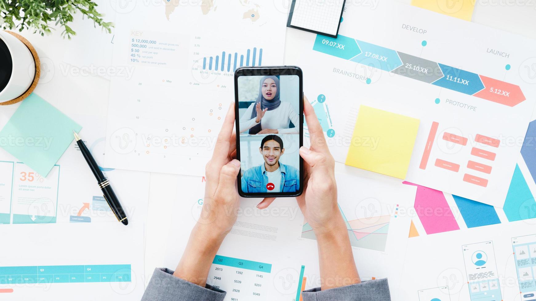 Vista superior de la joven empresaria asiática que usa el teléfono para hablar con un colega sobre el plan en la reunión de videollamada mientras trabaja desde casa en la sala de estar. distanciamiento social, cuarentena para la prevención del coronavirus. foto