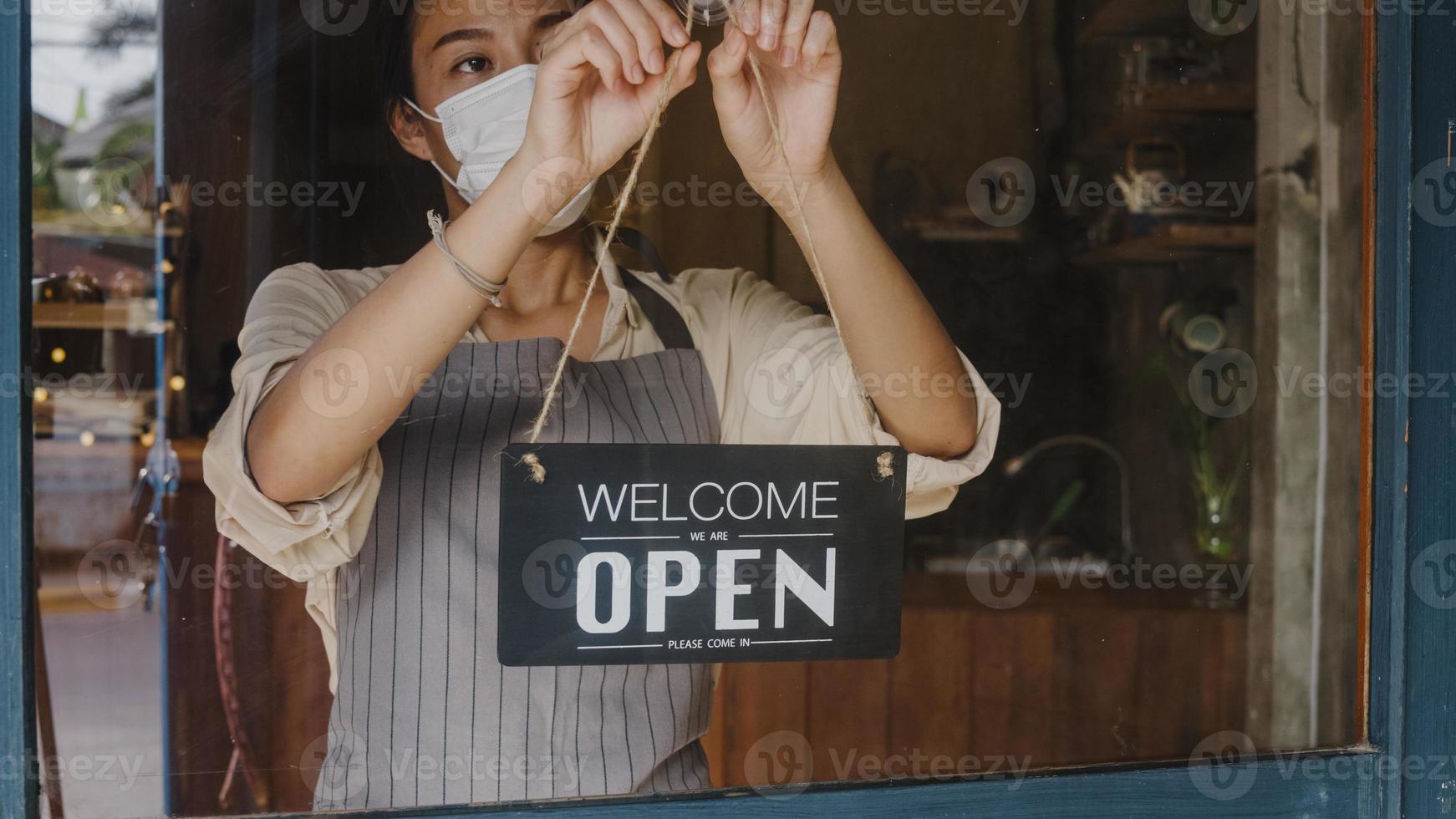 Young Asia girl wear face mask turning a sign from closed to open sign on door looking outside waiting for clients after lockdown. Owner small business, food and drink, business reopen again concept. photo